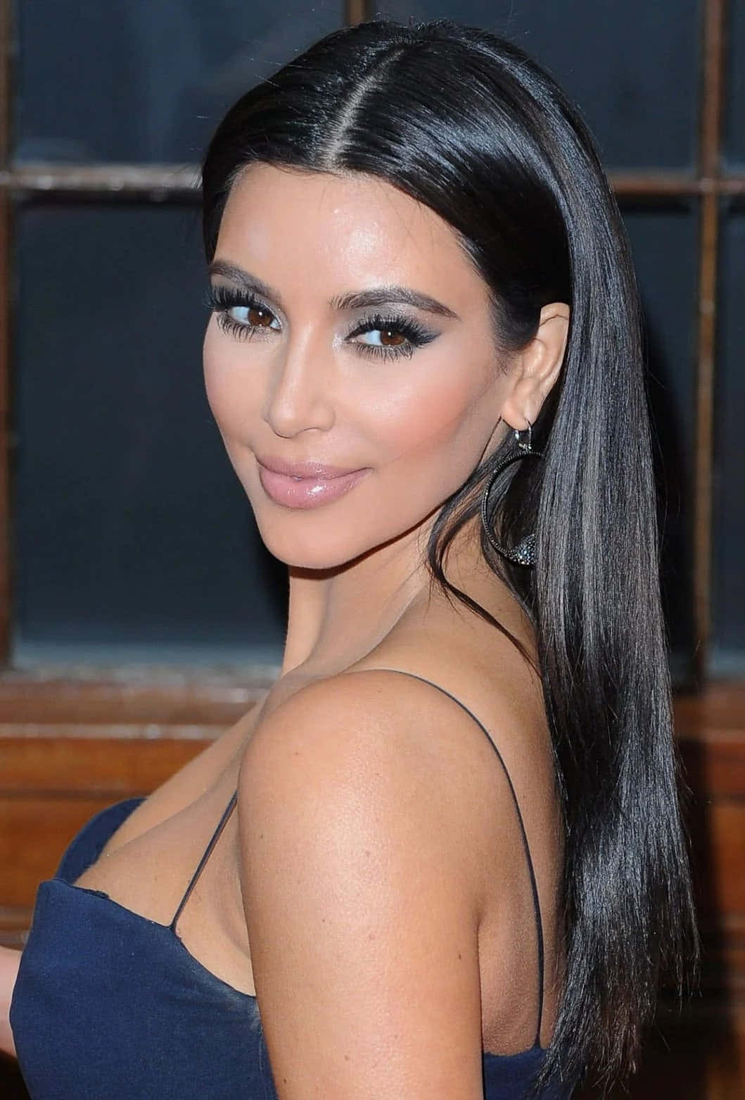 Kim Kardashian 桌布 壁紙 — 一位長黑髮、妝容大膽的迷人女性，身穿深藍色連衣裙，回頭微笑。