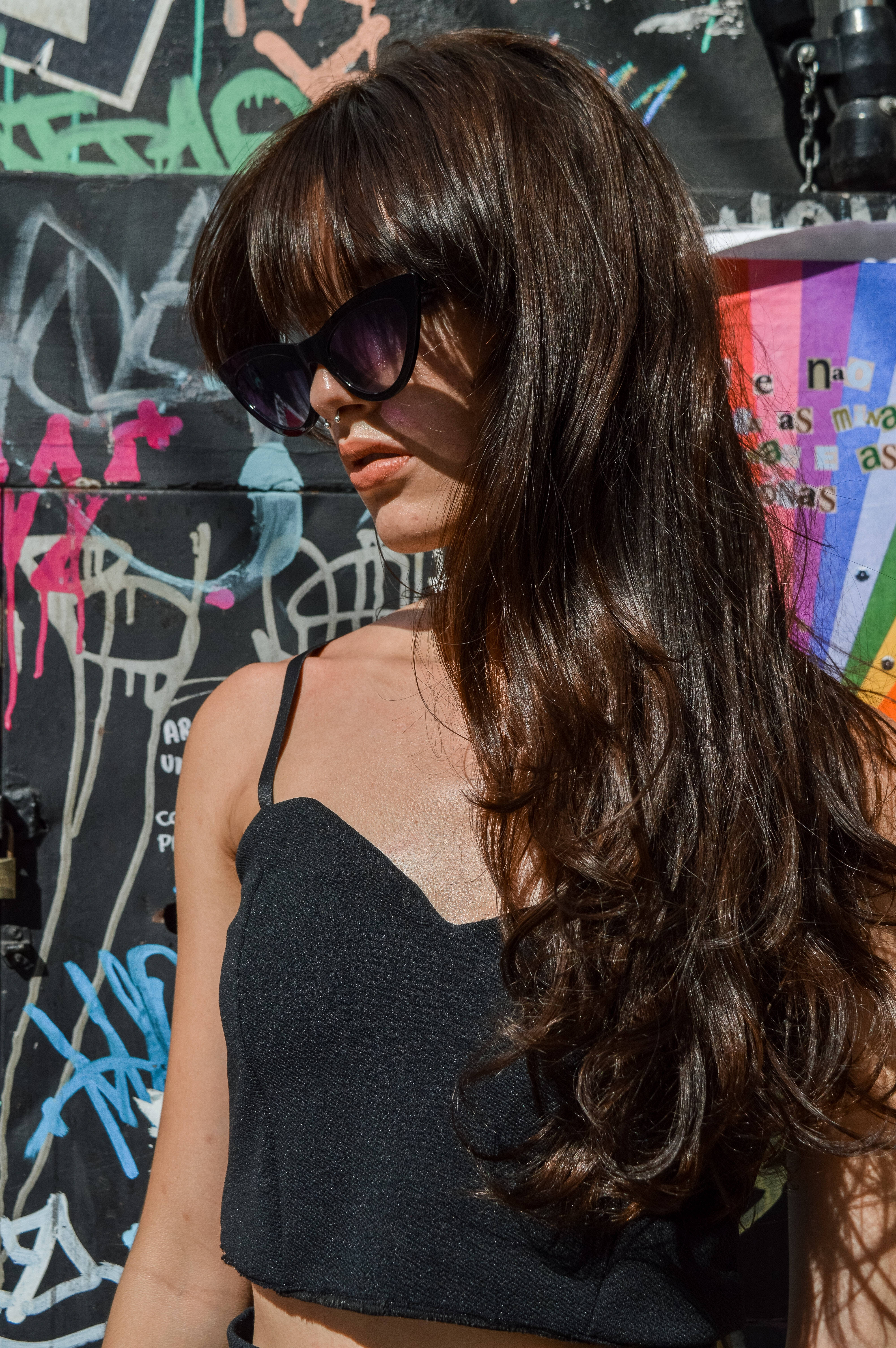 Sunglasses 桌布 壁紙 — 一位長黑髮、戴太陽眼鏡的女性，在明亮的陽光下站在充滿活力的塗鴉牆前。
