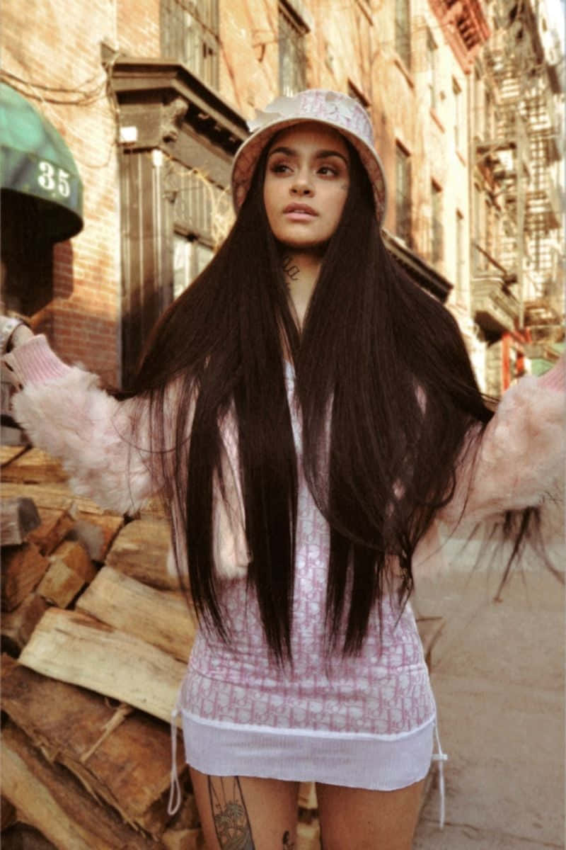kehlani 桌布 壁紙 — 一位長黑髮、戴漁夫帽的年輕女性站在陽光普照的城市小巷中，身穿粉紅色毛絨外套和印花迷你裙。