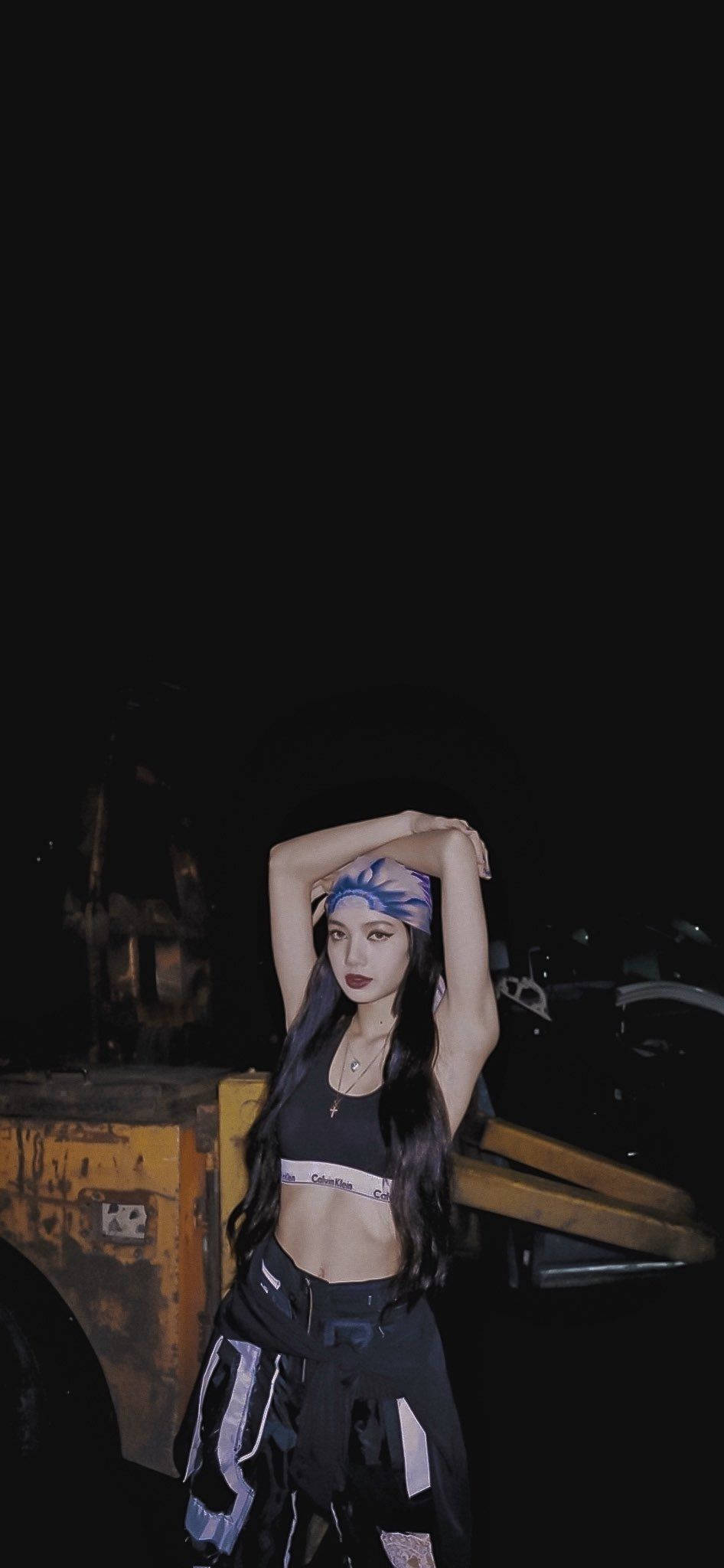 Lalisa 桌布 壁紙 — 一位長黑髮、戴著圖案頭帶的女性在夜間自信地擺姿勢，背景是黑暗的工業環境和一輛黃色車輛。