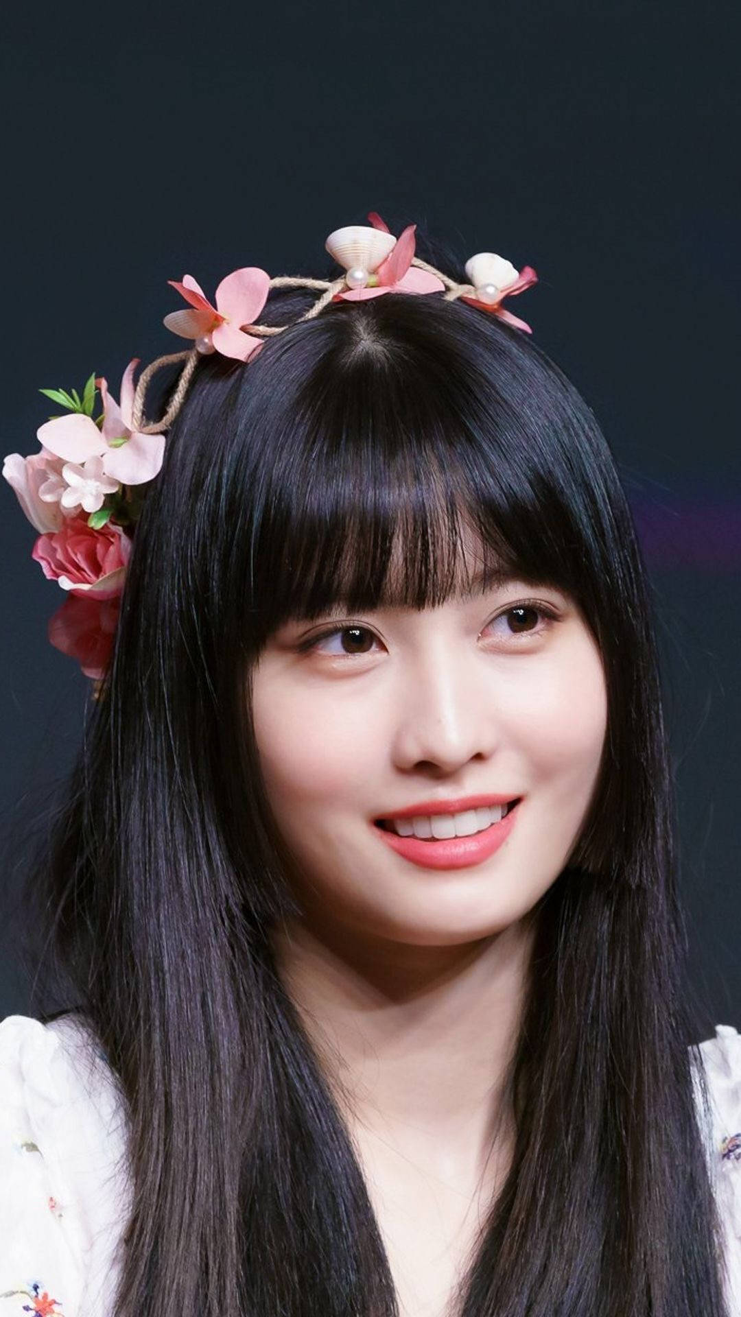 Momo 桌布 壁紙 — 一位長黑髮、戴著花卉頭飾的年輕女性在深色背景前溫暖微笑。