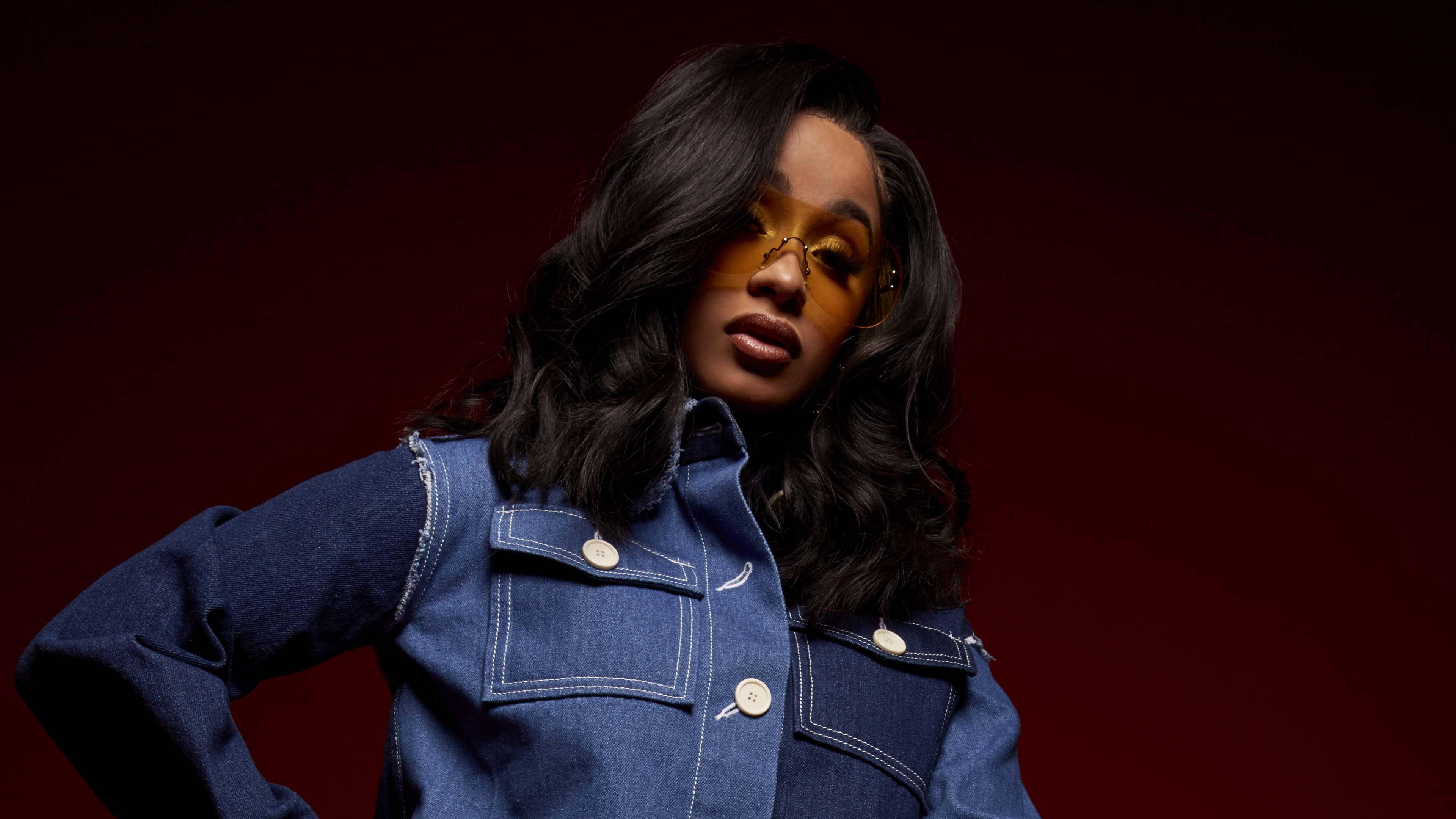 cardi 桌布 壁紙 — 一位長黑髮、戴黃色眼鏡的女性，身穿牛仔外套，在深紅色背景前自信地擺姿勢。
