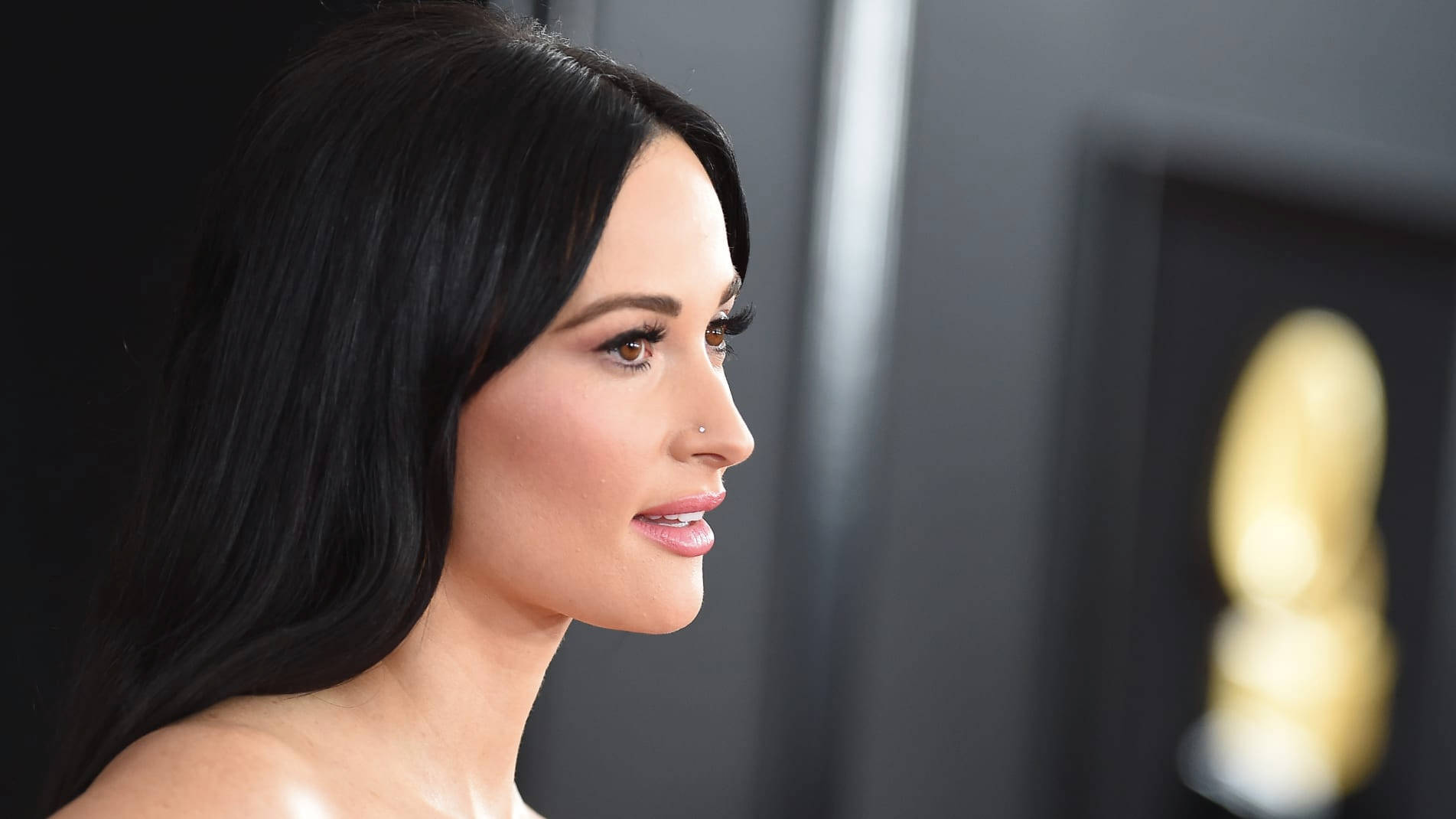 Kacey Musgraves 桌布 壁紙 — 一位長黑髮、有鼻環的女子的特寫側面，望向模糊的金色葛萊美獎座。