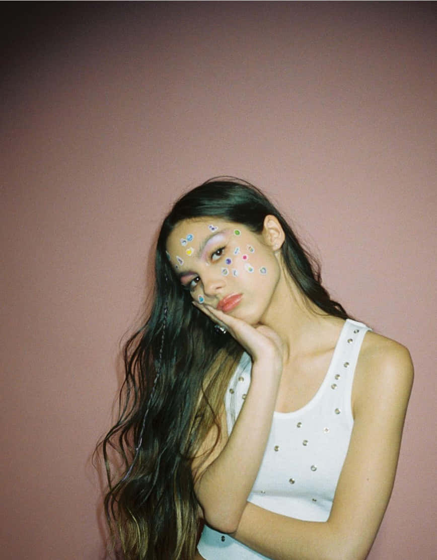 Olivia Rodrigo Aesthetic 桌布 壁紙 — 一位長黑髮、臉上貼著彩色貼紙的年輕女子，在柔和的粉紅色牆壁前若有所思地擺姿勢。