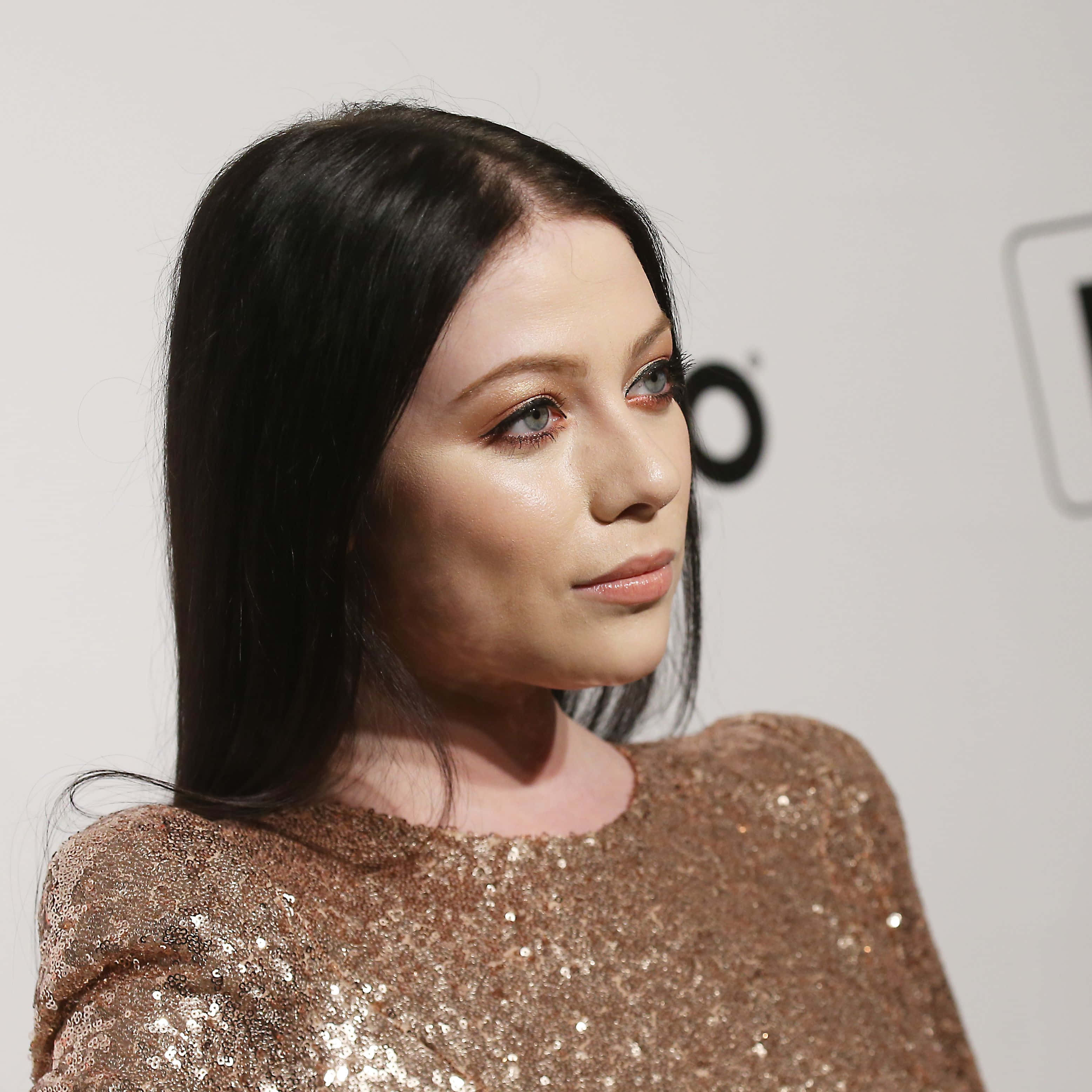 michelle trachtenberg 桌布 壁紙 — 一位長黑髮、藍眼睛引人注目的女性，身穿閃亮的玫瑰金亮片上衣，背景為純白色。