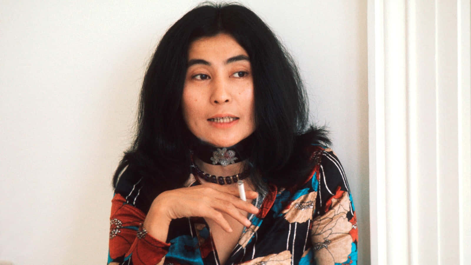 Smoking 桌布 壁紙 — 一位長黑髮、身穿圖案襯衫的女性，在純白牆壁前手持香煙，直視鏡頭。