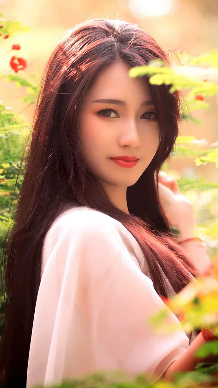 美女 桌布 壁紙 — 一位長黑髮、身穿白色上衣的女性站在陽光普照的綠植和紅花之間，溫柔地凝視著鏡頭。
