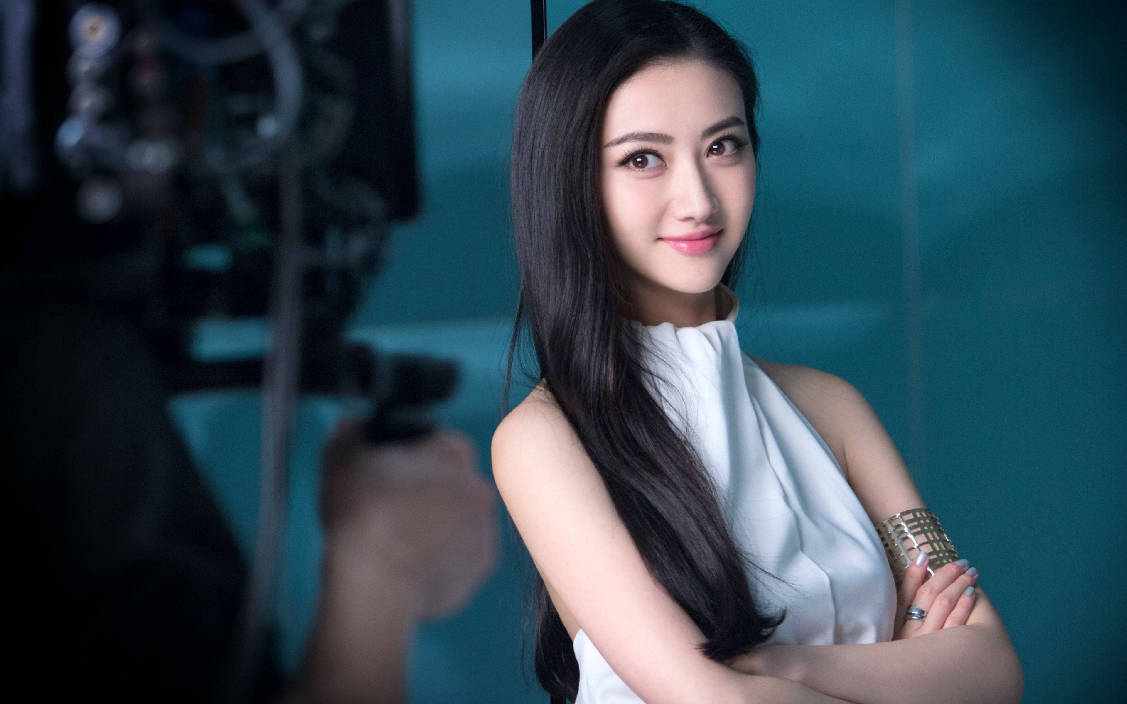 美女 桌布 壁紙 — 一位長黑髮、身穿白色連衣裙的優雅女性，在青綠色工作室背景前自信地交叉雙臂站立。