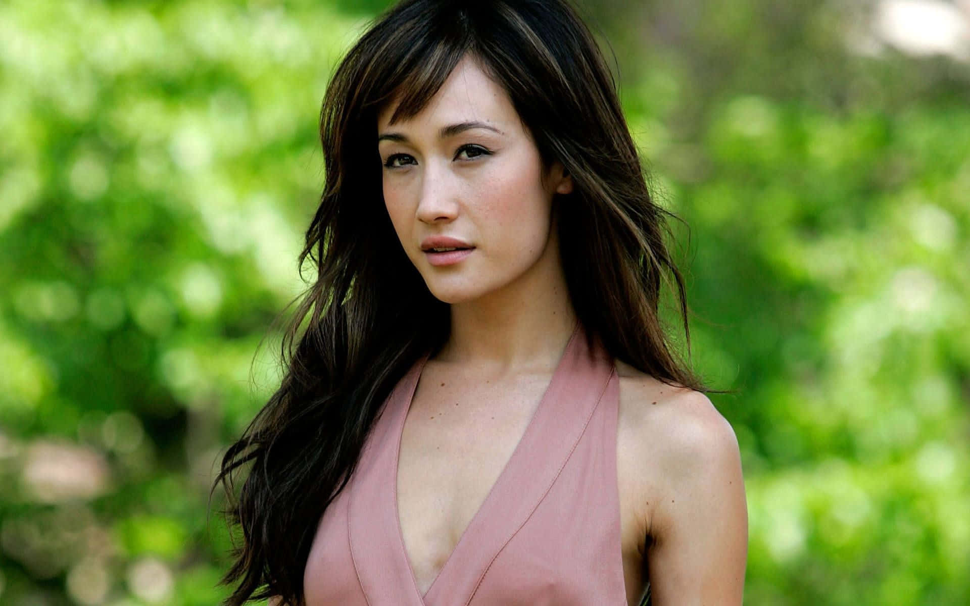 maggie q 桌布 壁紙 — 一位長黑髮、身穿粉紅色上衣的女性，站在光線柔和、綠意盎然的森林中。
