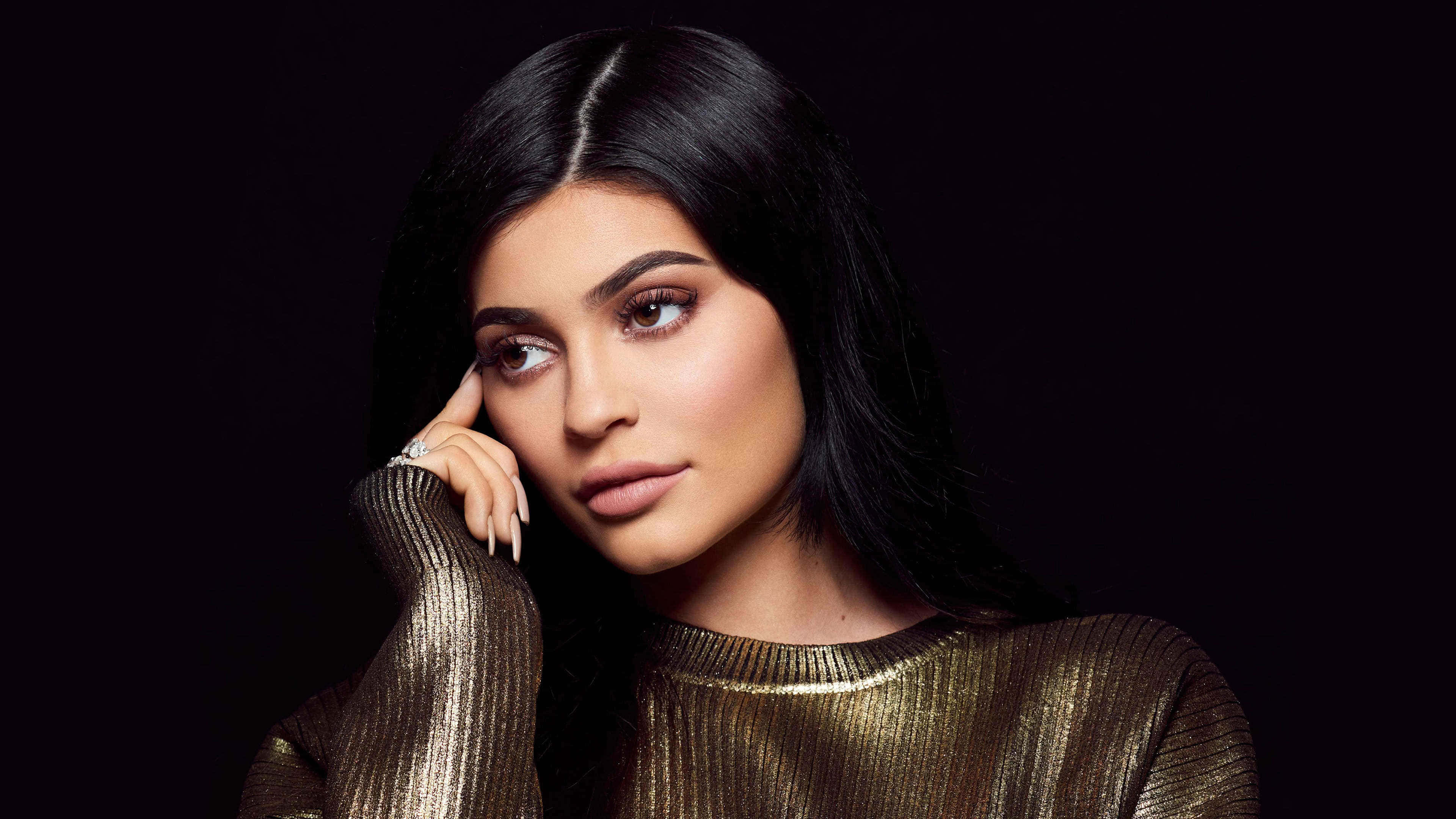 Kylie Jenner 4K 桌布 壁紙 — 一位長黑髮、身穿金色羅紋上衣的女性，在深色背景下若有所思地凝視著。