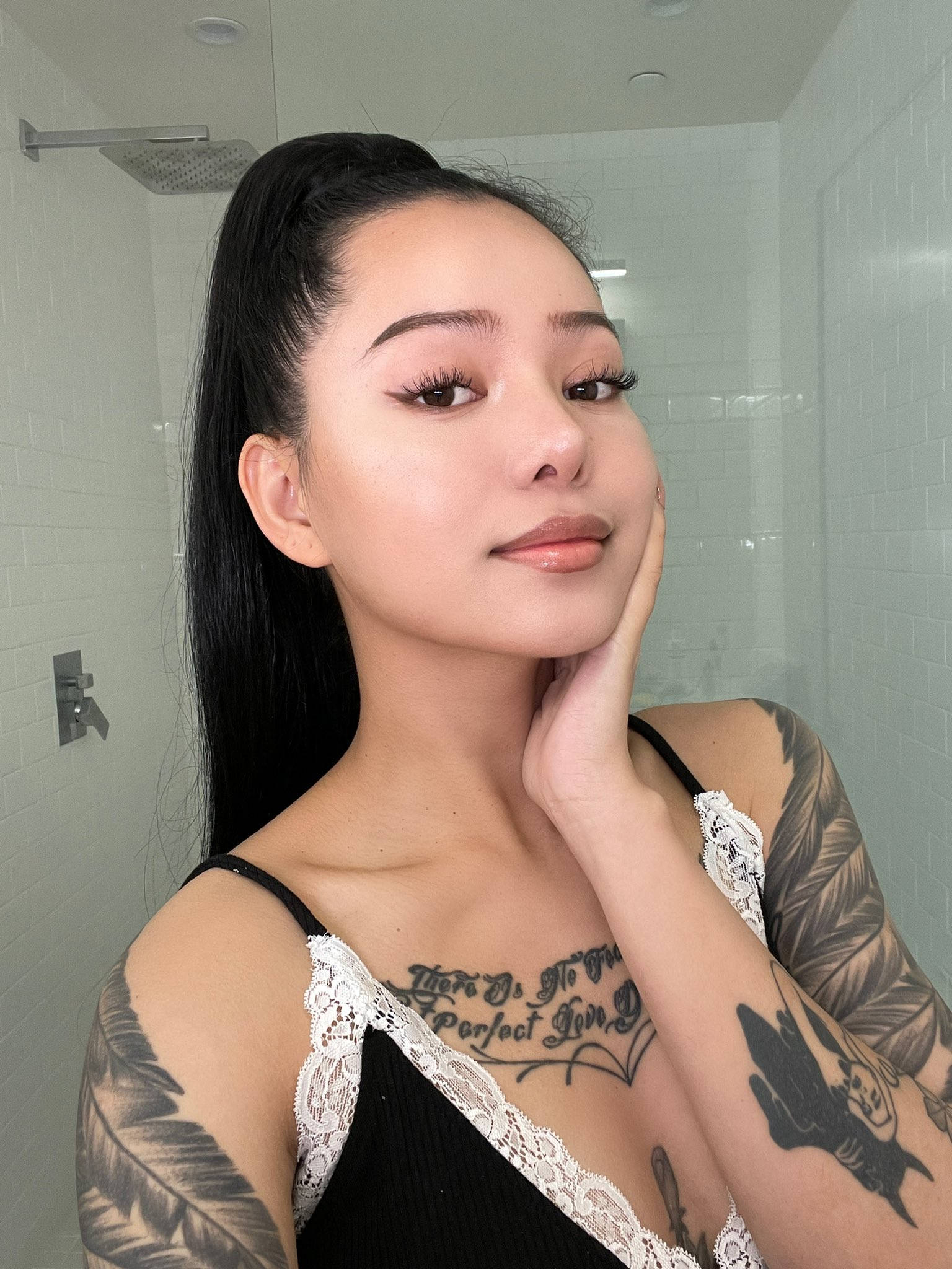 Bella Poarch 桌布 壁紙 — 一位長黑髮及有刺青的女性在白色磁磚浴室中穿著黑色蕾絲上衣擺姿勢。