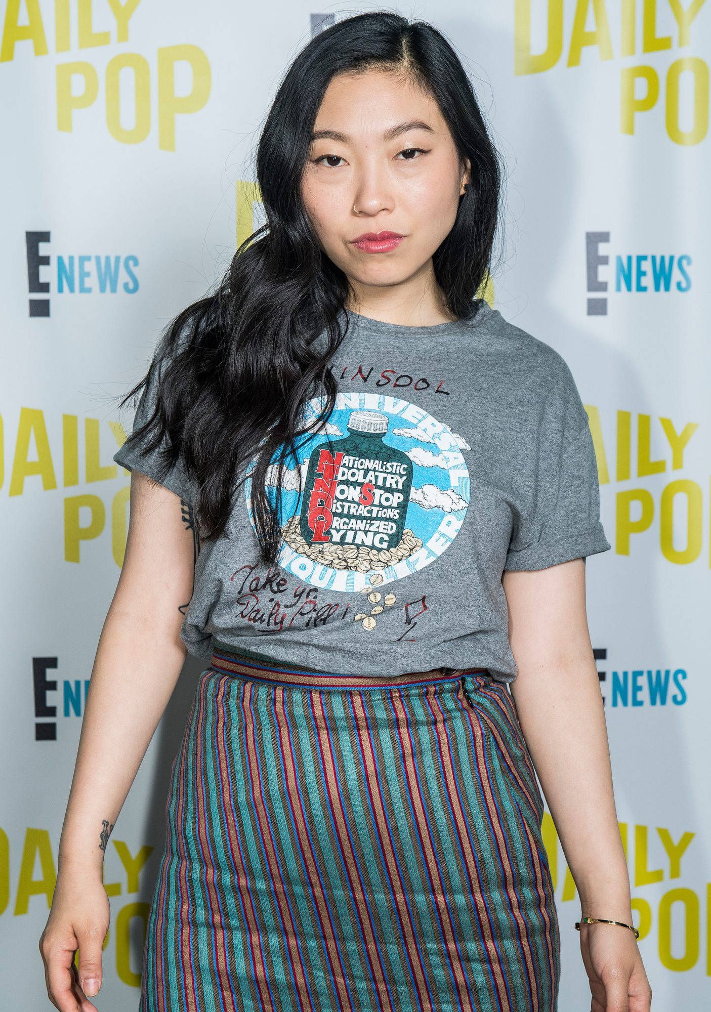 Awkwafina 桌布 壁紙 — 一位長黑髮女性穿著圖案T恤和條紋裙，背景為品牌活動場景。