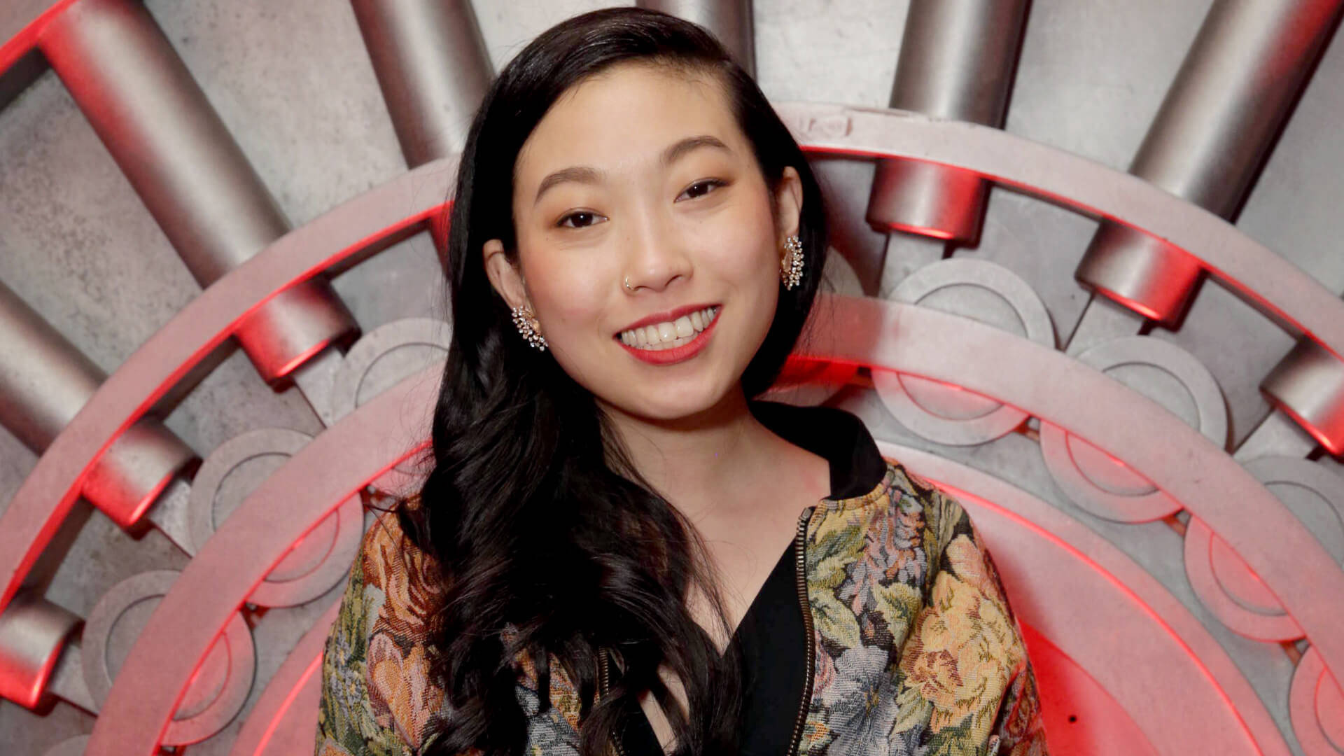 Awkwafina 桌布 壁紙 — 一位長黑髮微笑的女性，穿著花卉圖案外套，背景是紅色照明的金屬圓形結構。