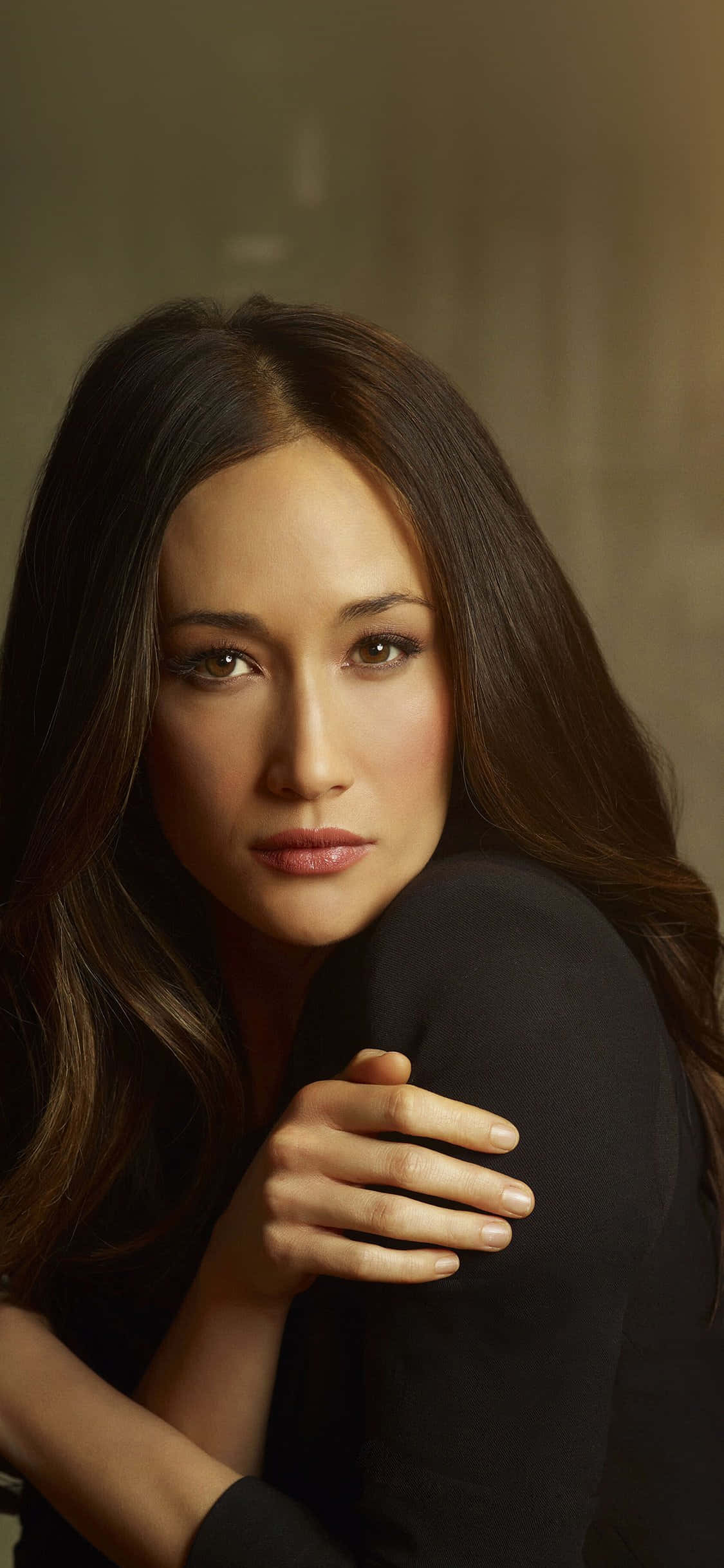 maggie q 桌布 壁紙 — 一位長黑髮的女性，在柔和的棕色背景上，將頭靠在手臂上，溫柔地凝視著。