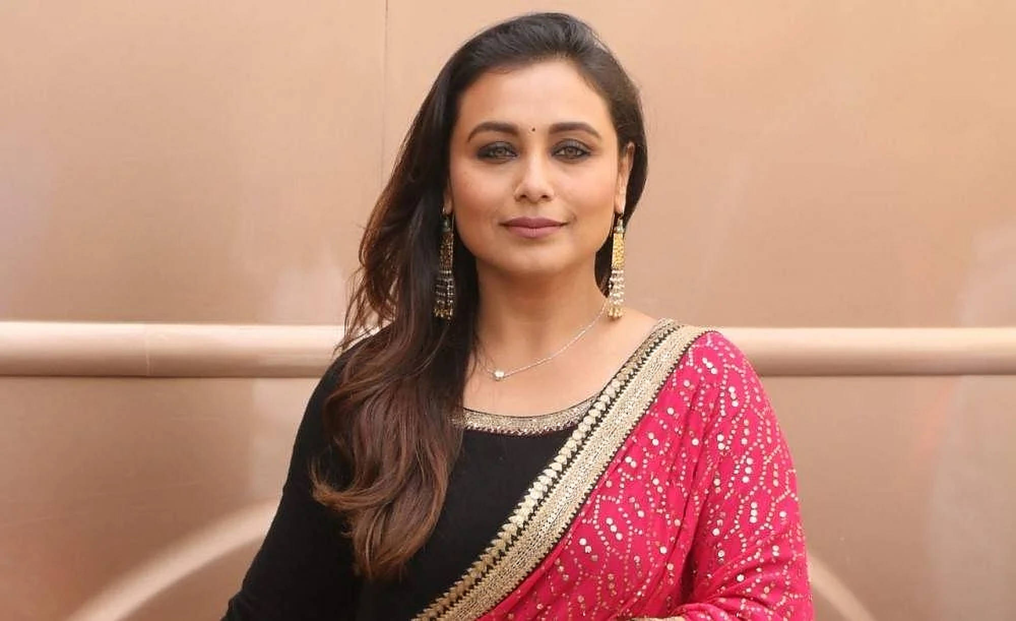 Rani Mukerji 桌布 壁紙 — 一位長黑髮的女性，在米色背景前穿著鮮豔的粉紅色和黑色傳統服飾，飾有金色刺繡和垂墜耳環。