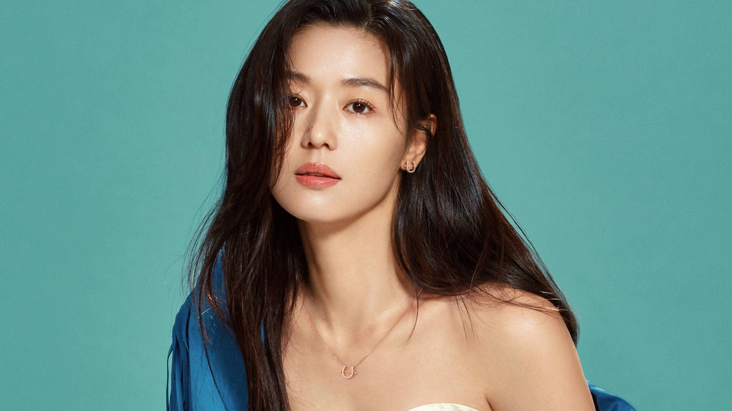 Jun Ji Hyun 桌布 壁紙 — 一位長黑髮的女性，在純青色背景前，穿著藍色垂墜上衣和精緻的金飾，溫柔地凝視著。