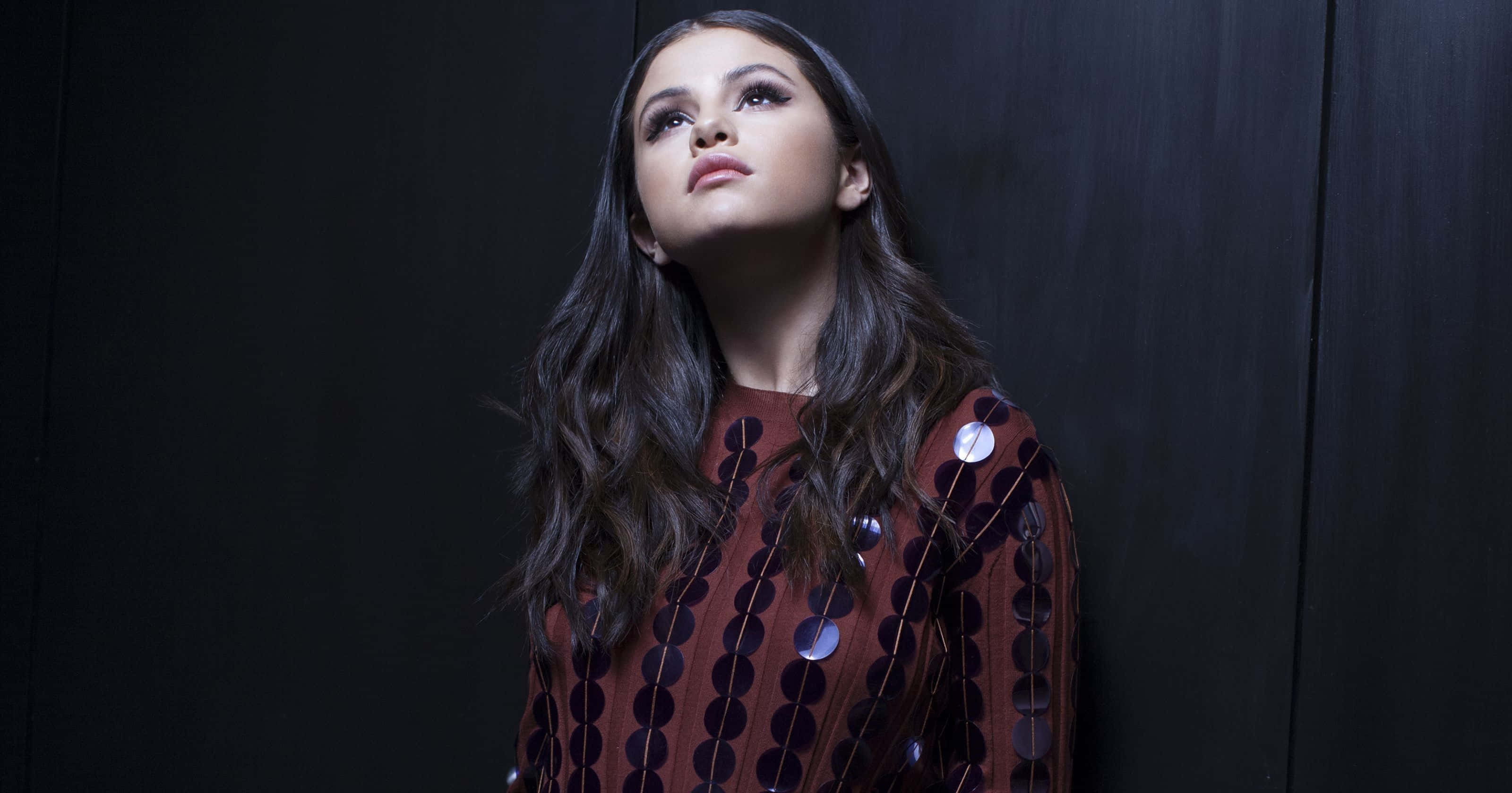 Selena Gomez 桌布 壁紙 — 一位長黑髮的女性，穿著帶有銀色圓片的紅色圖案連衣裙，在深色木質背景下向上凝視。