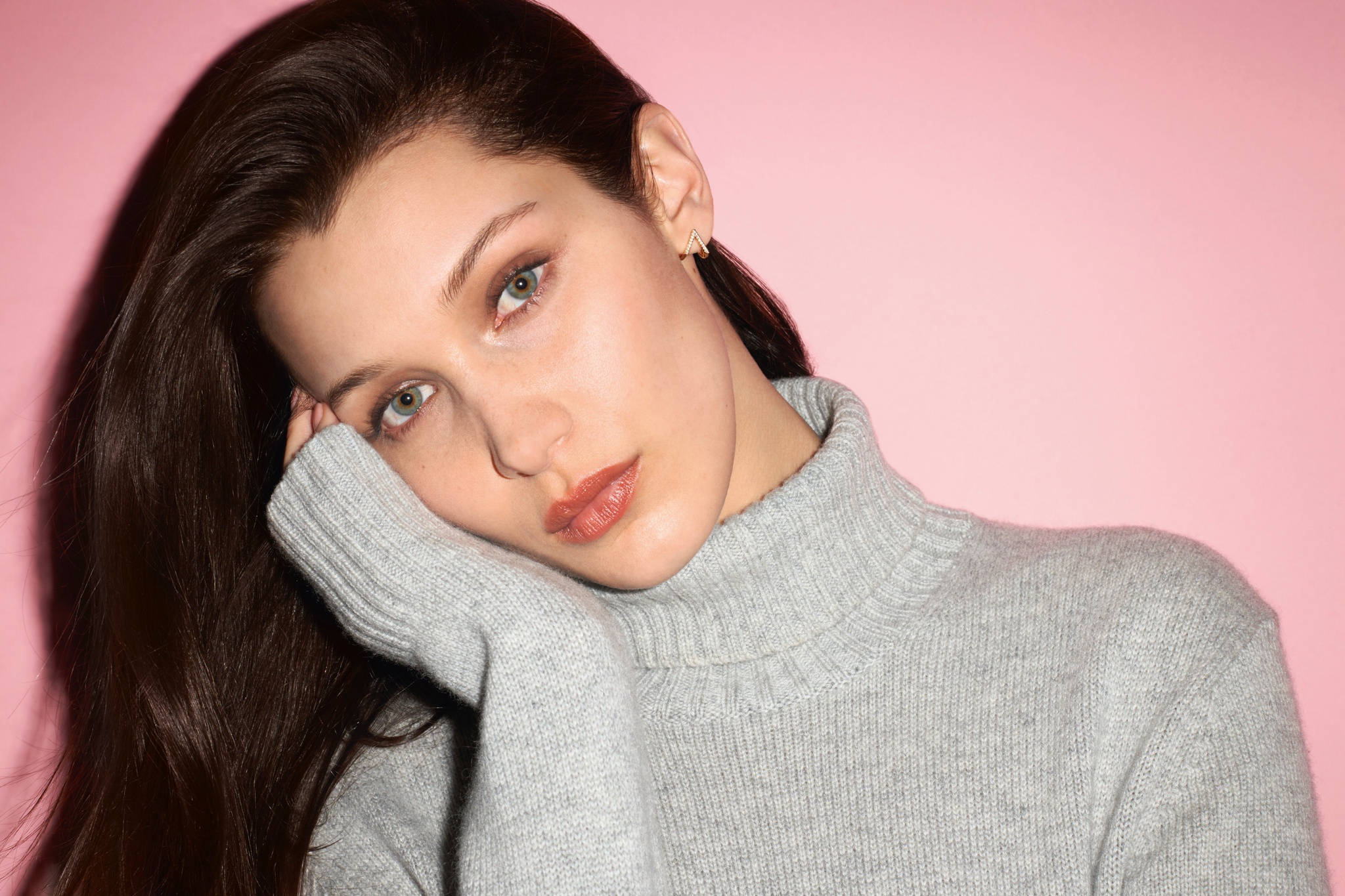 Bella Hadid 桌布 壁紙 — 一位長黑髮的女性，穿著灰色高領毛衣，在柔和的粉紅色背景上將頭靠在手上。