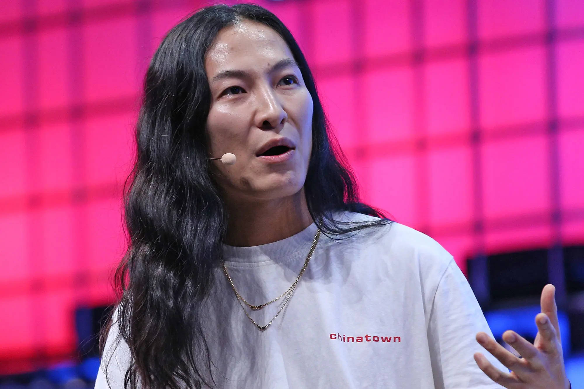 Alexander Wang 桌布 壁紙 — 一位長黑髮的女性在充滿活力的粉紅色網格背景前熱情地在舞台上發言。