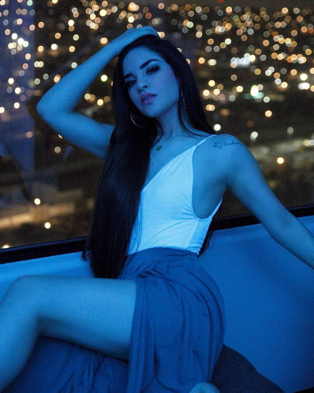 美學 桌布 壁紙 — 一位長黑髮的女性在夜晚坐在窗邊，沐浴在冷藍光中，若有所思地凝視著，身後的城市燈光模糊成散景。