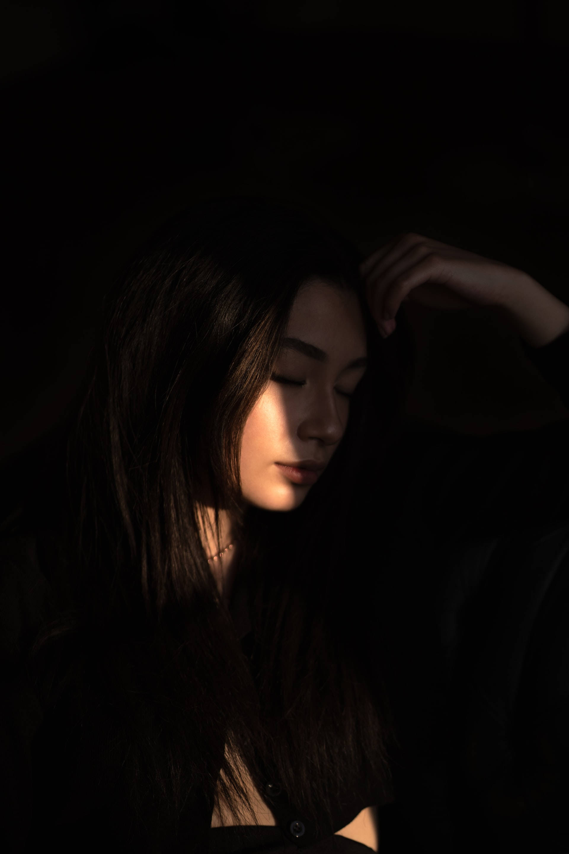 女孩 桌布 壁紙 — 一位長黑髮的女性在昏暗的空間中將手放在頭上，沐浴在柔和側光中，突顯她平靜的表情。
