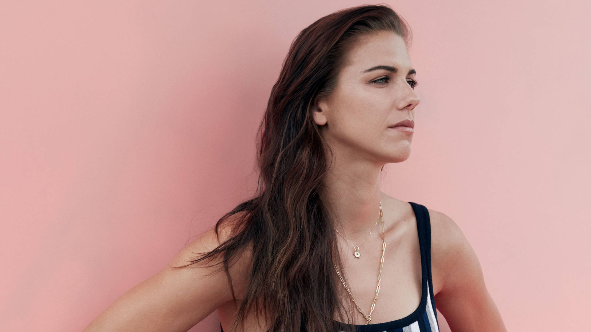 Alex Morgan 桌布 壁紙 — 一位長黑髮的女性在柔和的粉紅色工作室背景下若有所思地凝視，穿著條紋上衣和分層的金項鍊。
