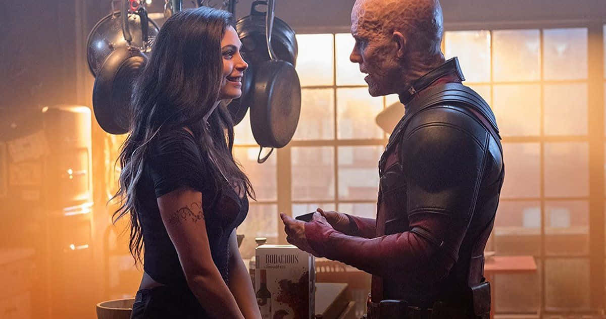 Deadpool And Vanessa 桌布 壁紙 — 一位長黑髮的女性在溫暖照明的廚房裡，掛著鍋具，對一位穿著皮衣的光頭男子溫暖地微笑。