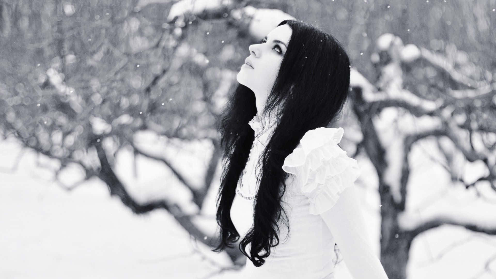 Winter Fashion 桌布 壁紙 — 一位長黑髮的女性在白雪覆蓋的冬季森林中向上凝望，周圍是光禿的樹枝和飄落的雪花。
