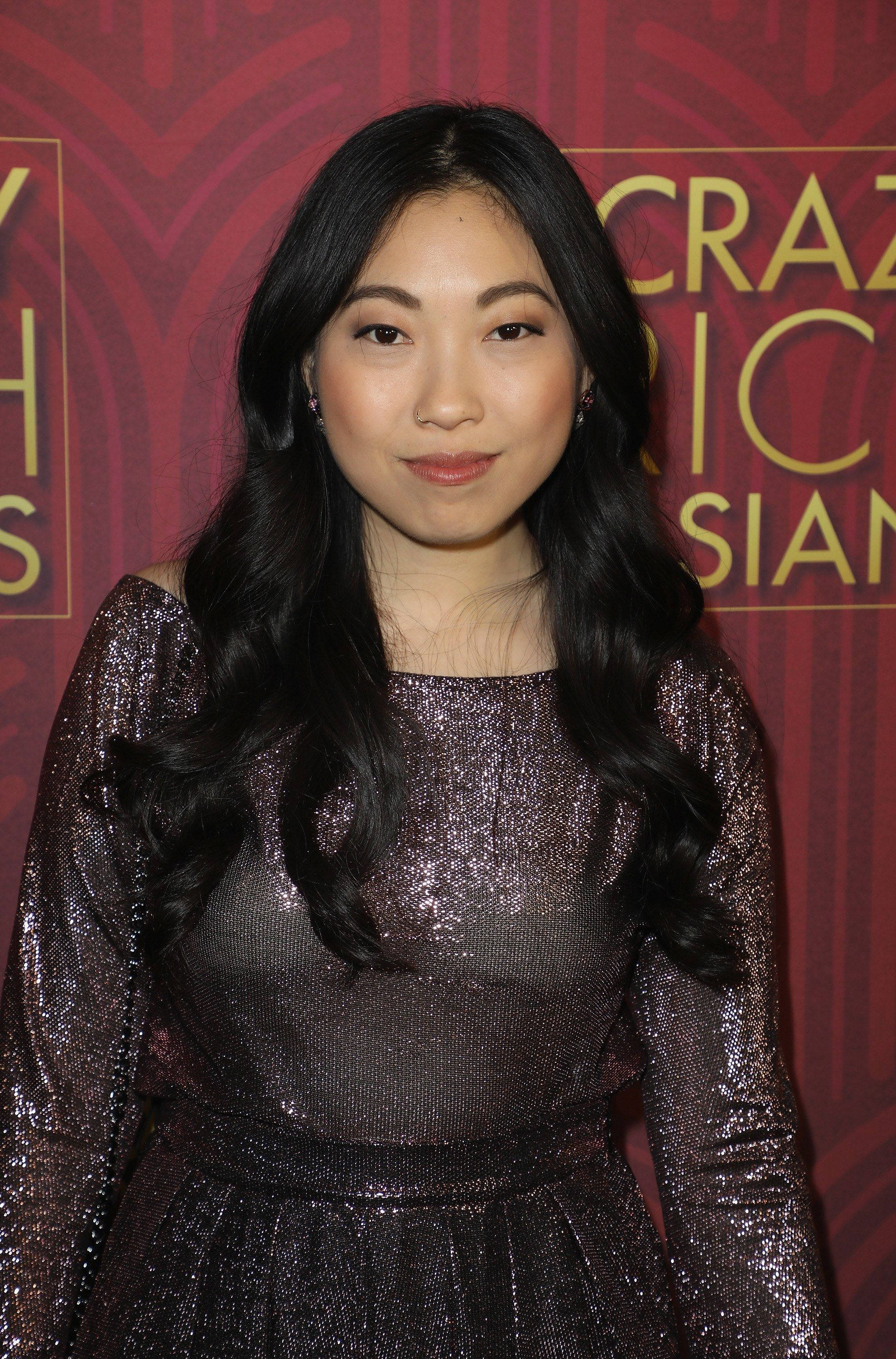 Awkwafina 桌布 壁紙 — 一位長黑髮的女性在紅地毯活動中穿著閃亮的紫色連衣裙，背景為金色文字。