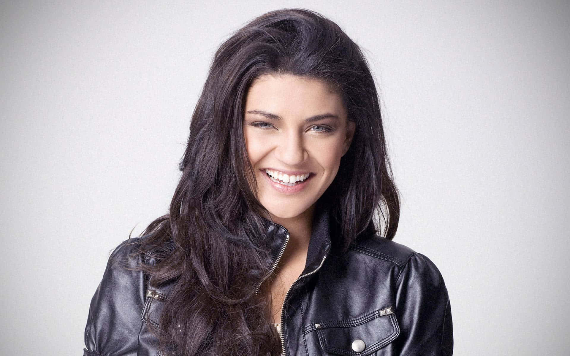 jessica szohr 桌布 壁紙 — 一位長黑髮的女性在純灰色背景前穿著黑色皮夾克，露出燦爛的笑容。