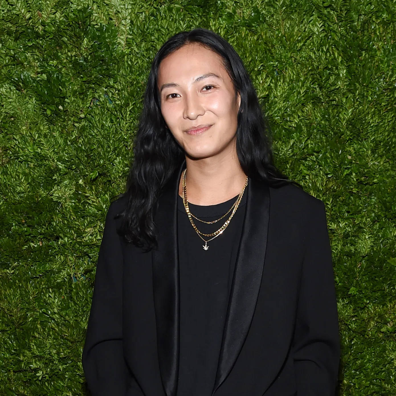 Alexander Wang 桌布 壁紙 — 一位長黑髮的女性在茂密的綠色樹籬牆前溫柔微笑，身穿黑色西裝外套和層疊的金色項鍊。