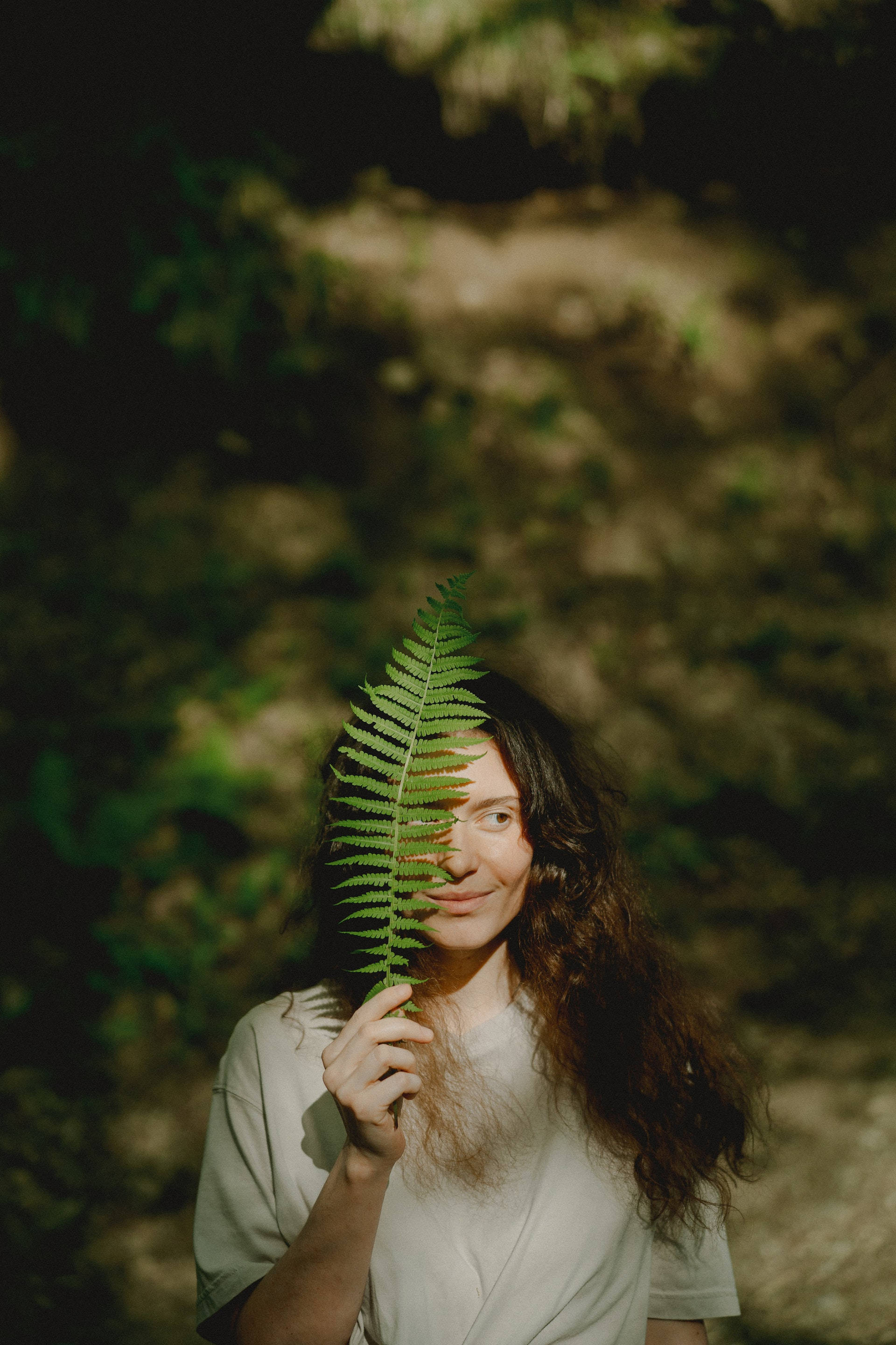 Plant Aesthetic 桌布 壁紙 — 一位長黑髮的女性在陽光斑駁的森林中，手持一片鮮綠的蕨葉遮住臉龐。