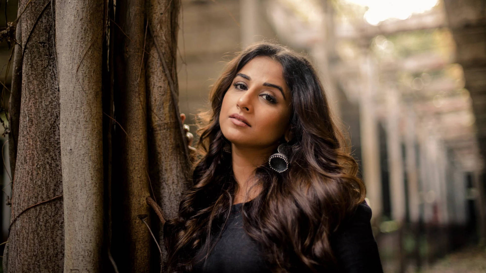 Vidya Balan 桌布 壁紙 — 一位長黑髮的女性在陽光普照的小巷中靠著樹幹，背景是模糊的建築物。