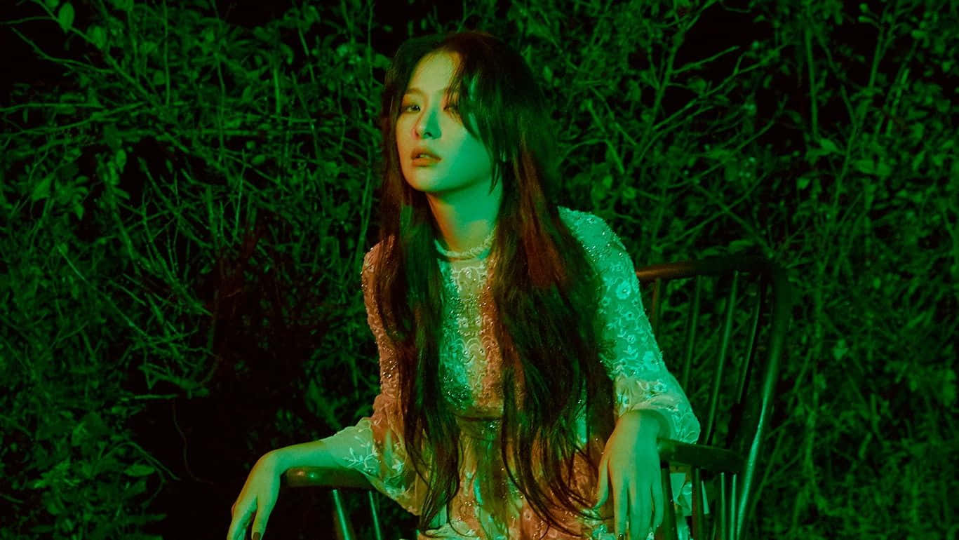 K-pop 桌布 壁紙 — 一位長黑髮的女性坐在椅子上，周圍是茂密的綠色樹葉，夜晚在詭異的綠色燈光下。