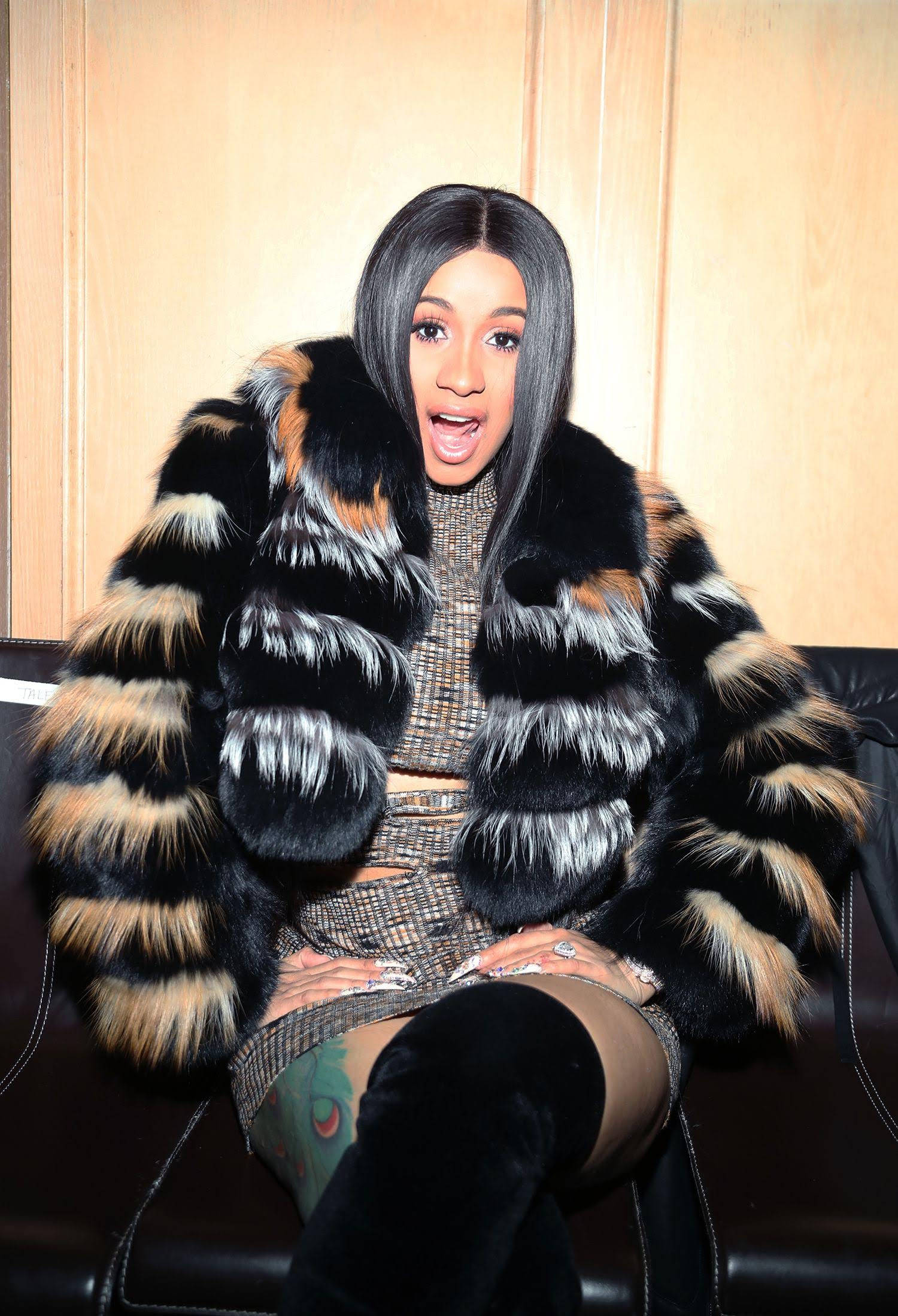 cardi 桌布 壁紙 — 一位長黑髮的女性坐在黑色皮革沙發上，穿著一件大膽的黑、棕、白毛皮大衣，內搭質感連衣裙。