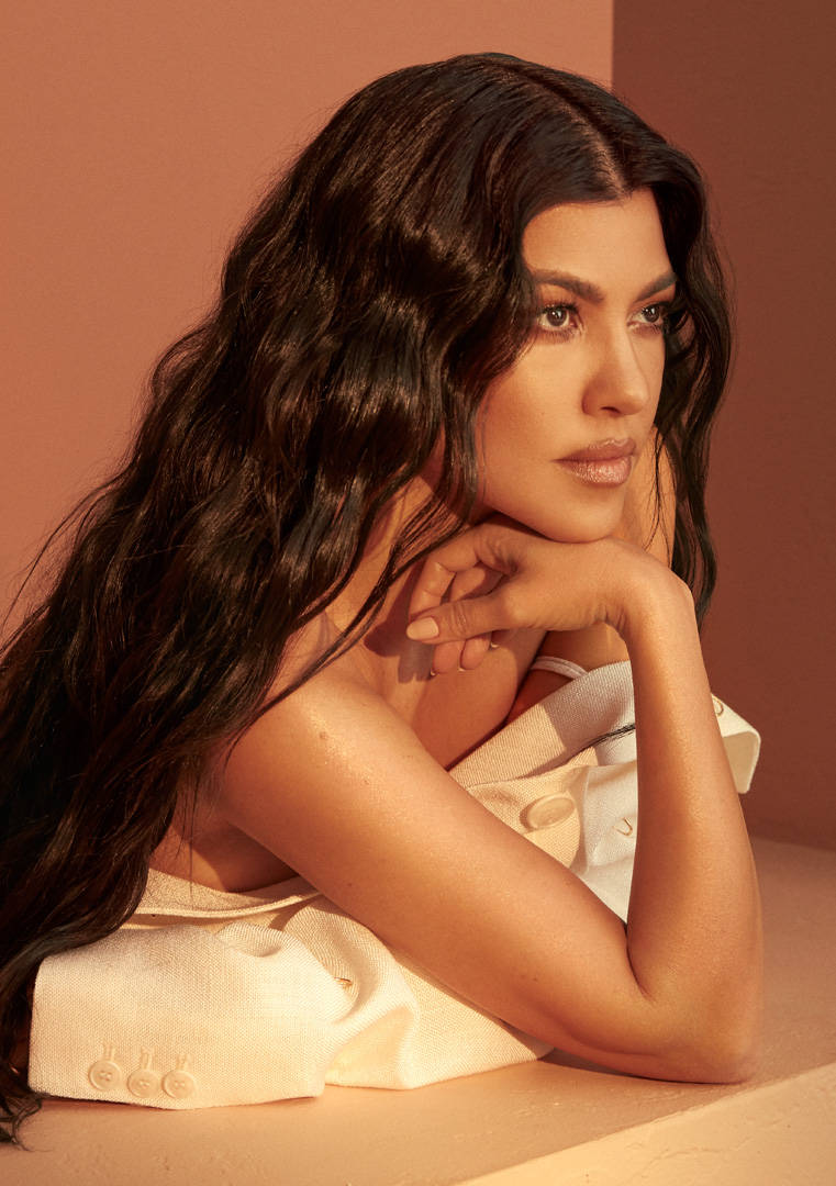 Kourtney 桌布 壁紙 — 一位長黑髮的女性將下巴擱在手上，靠著溫暖的赤陶牆，沐浴在柔和的金色光線中。