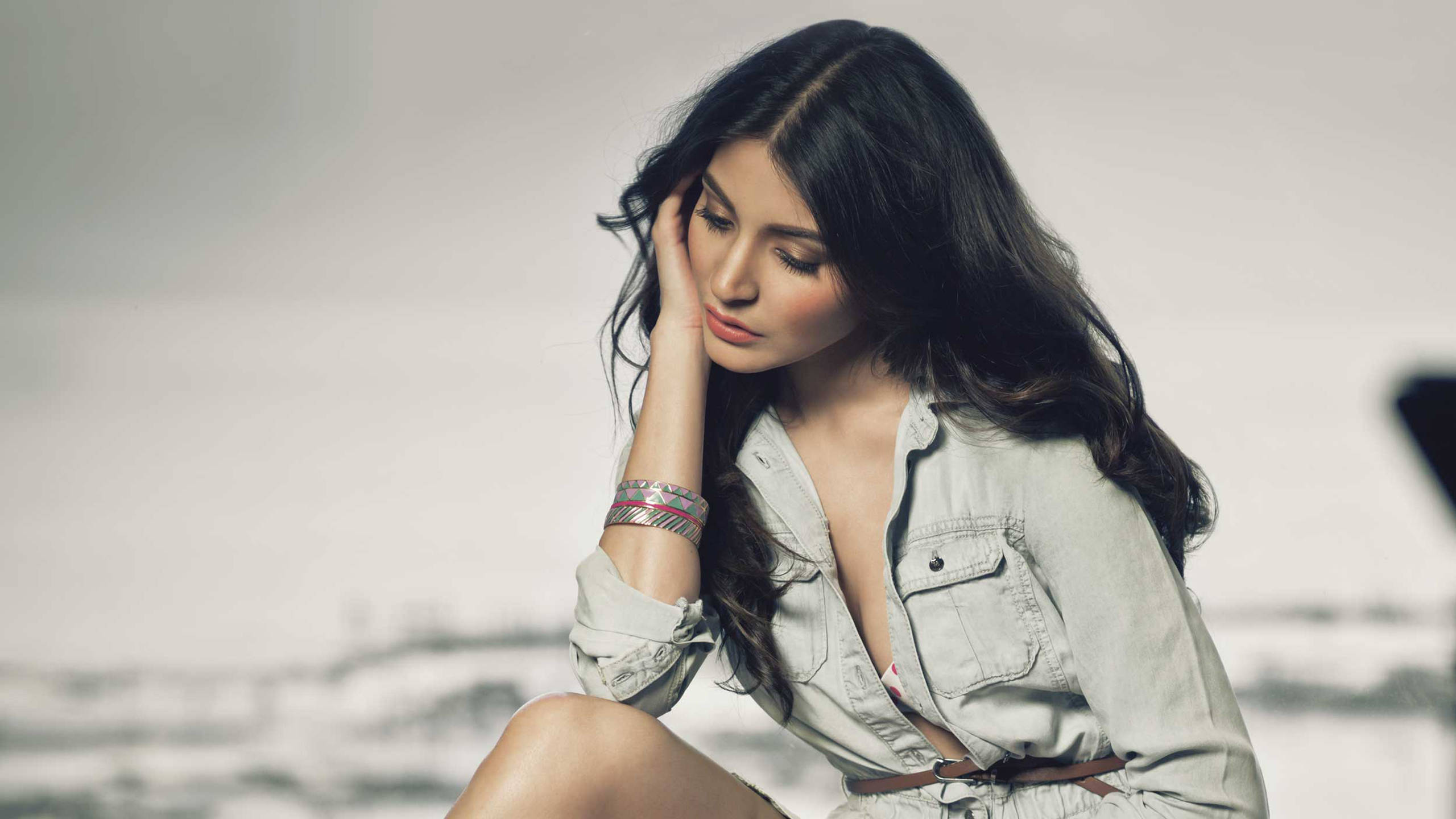 Anushka Sharma 桌布 壁紙 — 一位長黑髮的女性沉思地坐著，手托著臉頰，穿著淺灰色襯衫，背景柔和模糊。