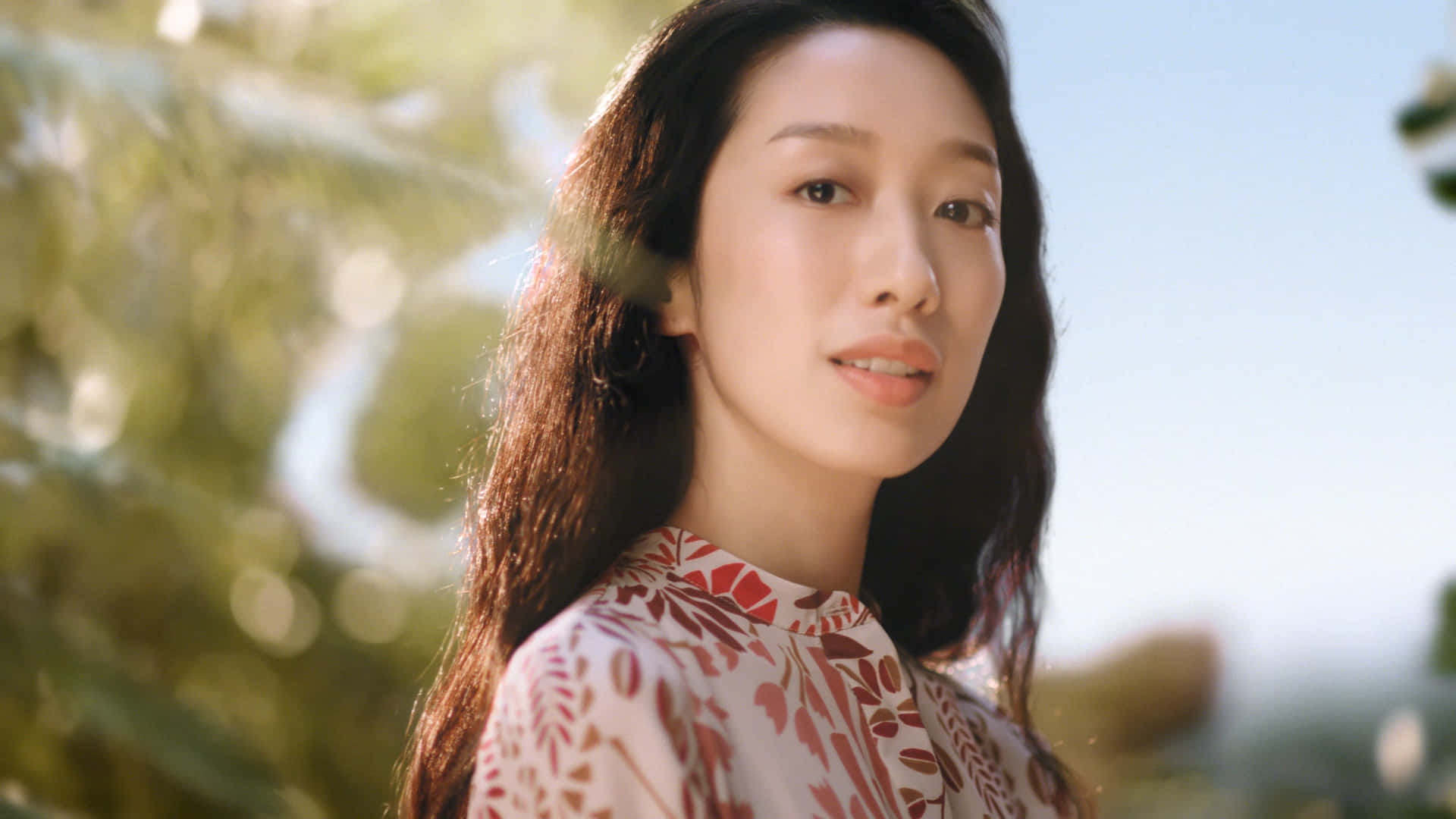 美女 桌布 壁紙 — 一位長黑髮的女性穿著花卉圖案上衣，在陽光普照的綠色背景下溫柔地凝視鏡頭。