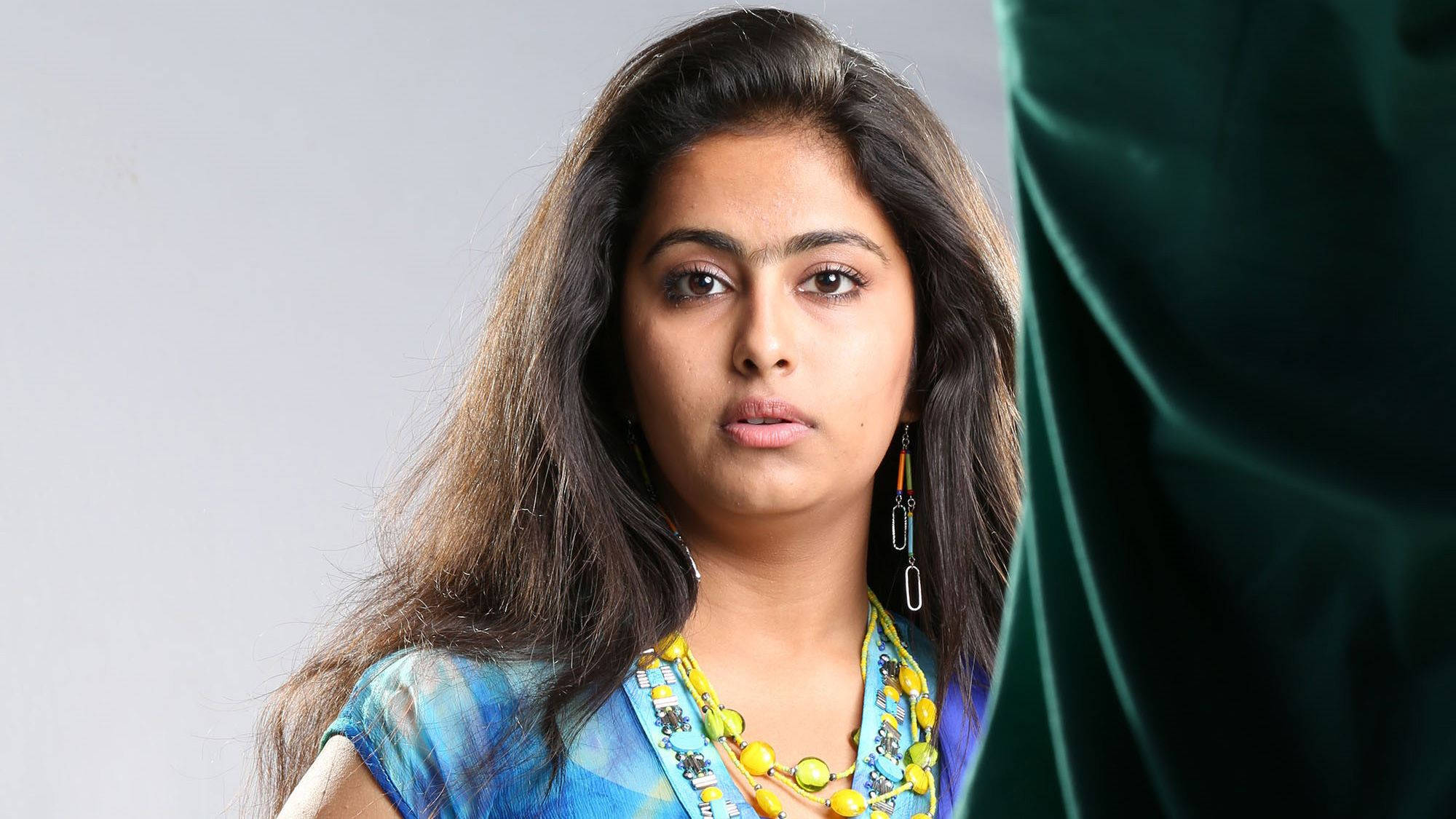 telugu 桌布 壁紙 — 一位長黑髮的女性穿著藍色圖案上衣和黃色珠鏈，在綠色天鵝絨簾幕的樸素背景下直視鏡頭。