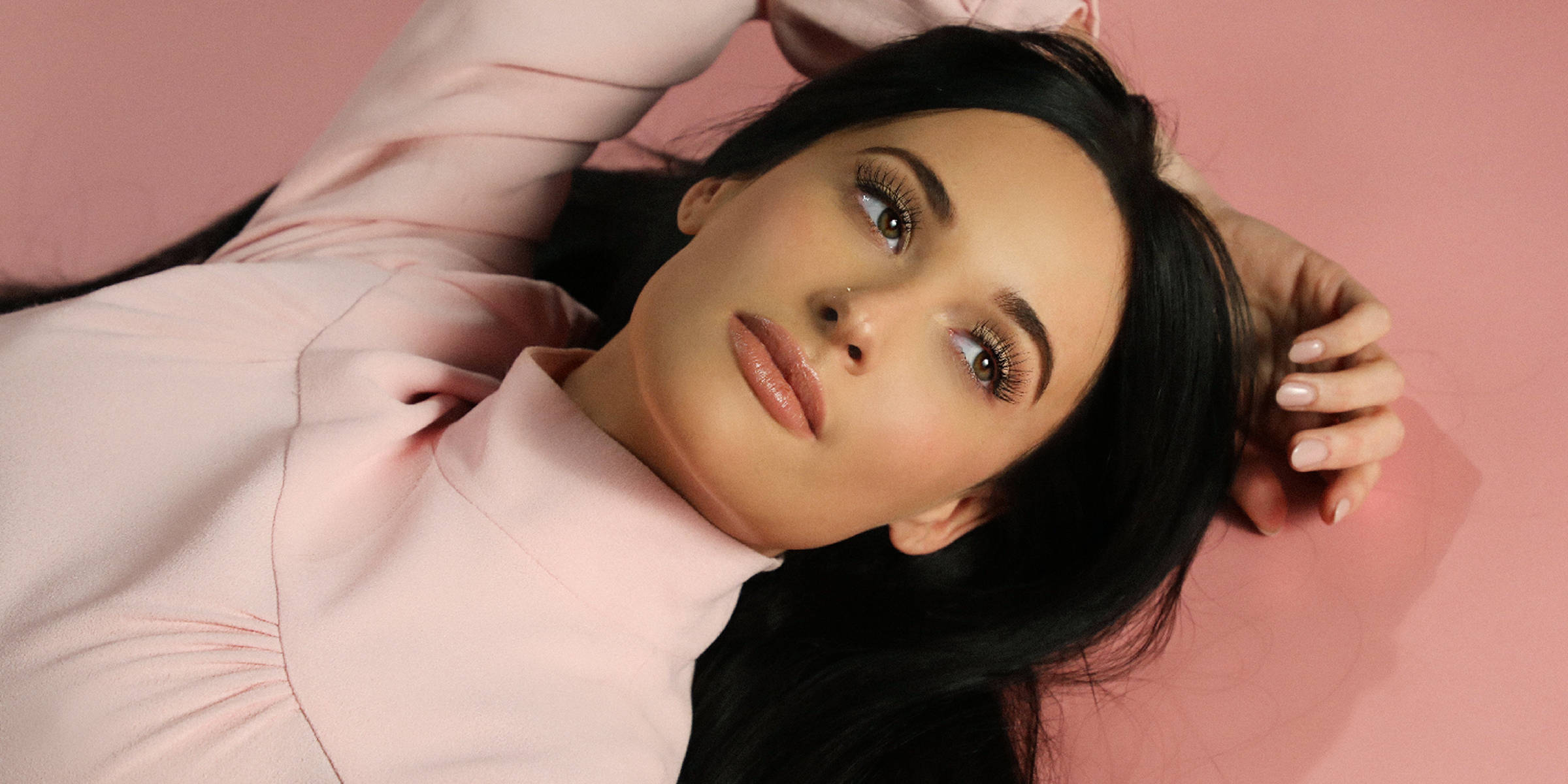 Kacey Musgraves 桌布 壁紙 — 一位長黑髮的女性躺在柔軟的粉紅色背景上，帶著平靜的表情向上凝視。