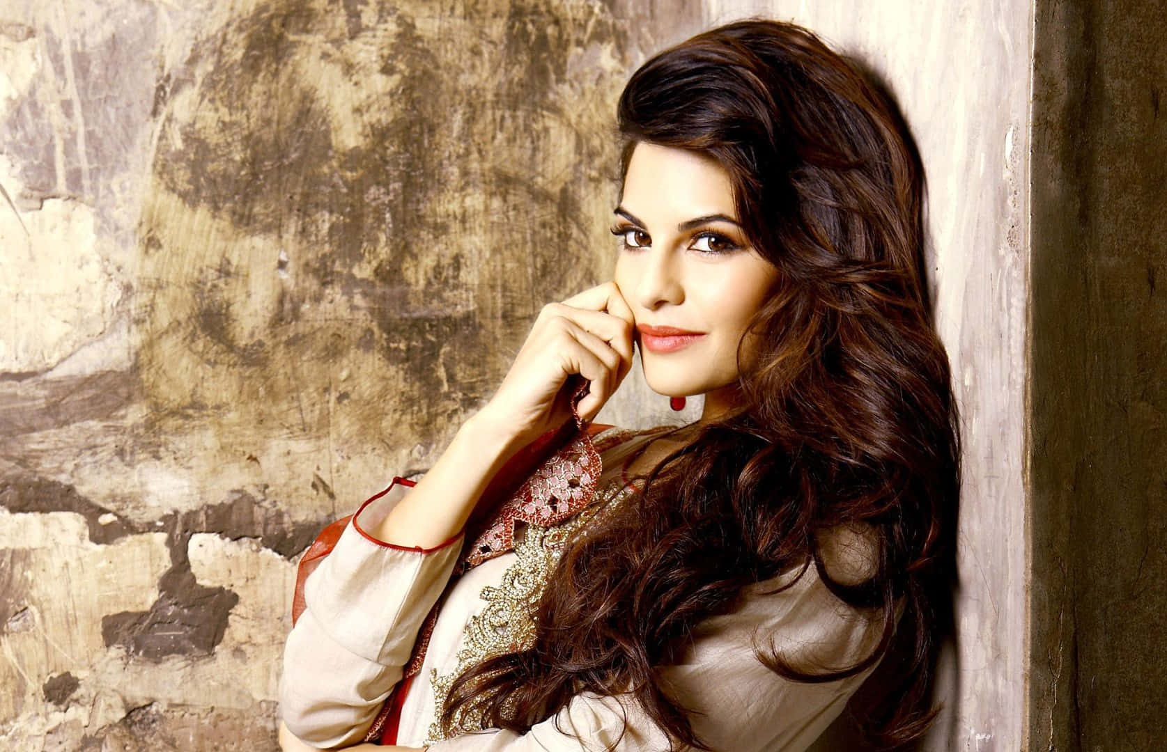 Jacqueline Fernandez 桌布 壁紙 — 一位長黑髮的女性靠著風化的牆壁，手靠近臉龐，穿著華麗的奶油色和紅色服裝。