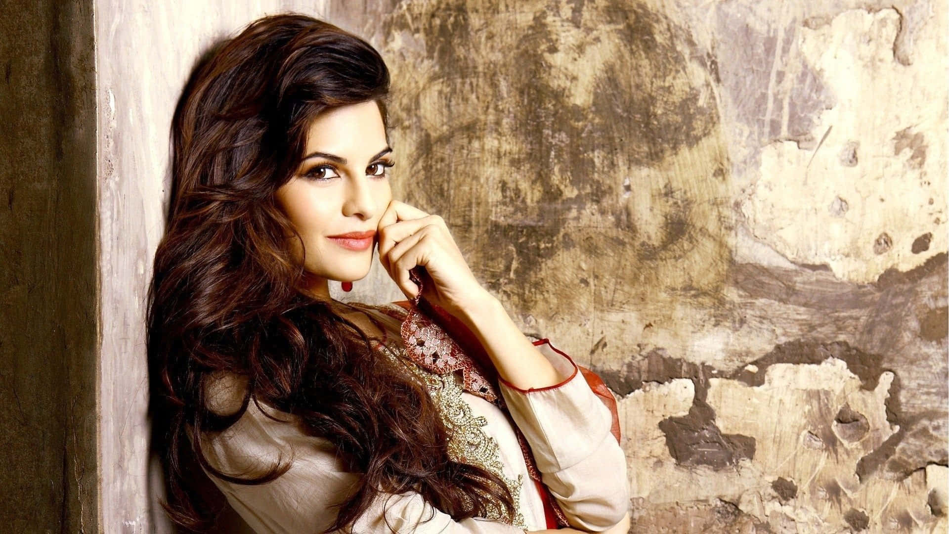 Jacqueline Fernandez 桌布 壁紙 — 一位長黑髮的女性靠著風化的牆壁，手靠近臉龐，在柔和光線的肖像中。