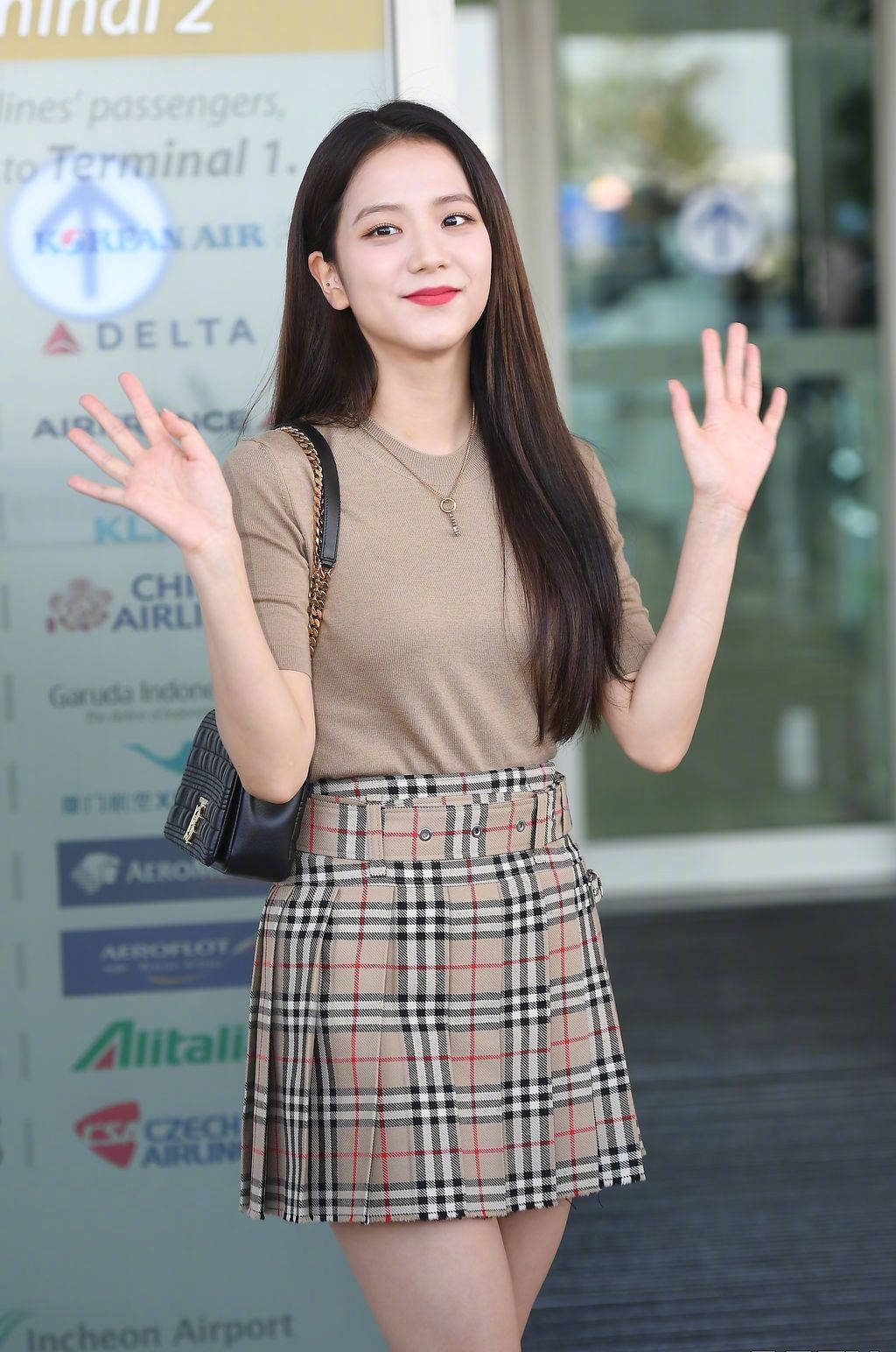 Burberry 桌布 壁紙 — 一位長黑髮的年輕女子在機場航站樓穿著米色上衣和格子裙，開心地揮手。