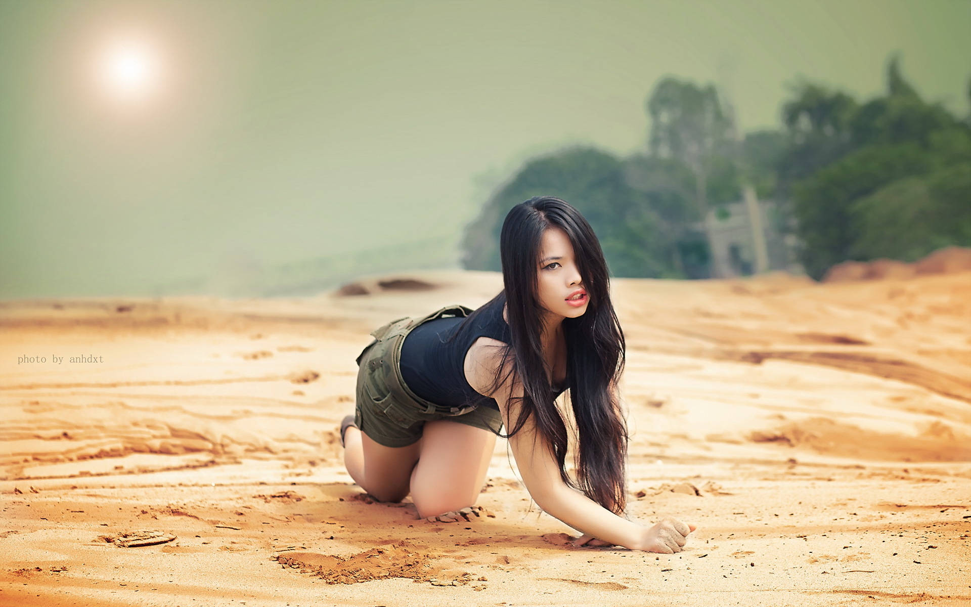 Asian Woman 桌布 壁紙 — 一位長黑髮的年輕女子跪在陽光照耀的沙地上，神情平靜地直視鏡頭。