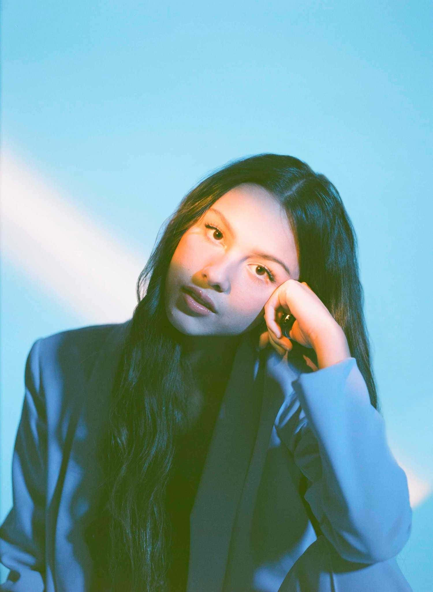 Olivia Rodrigo Aesthetic 桌布 壁紙 — 一位長黑髮的年輕女性，在清涼的藍色漸變背景上用手托著頭，光線柔和。