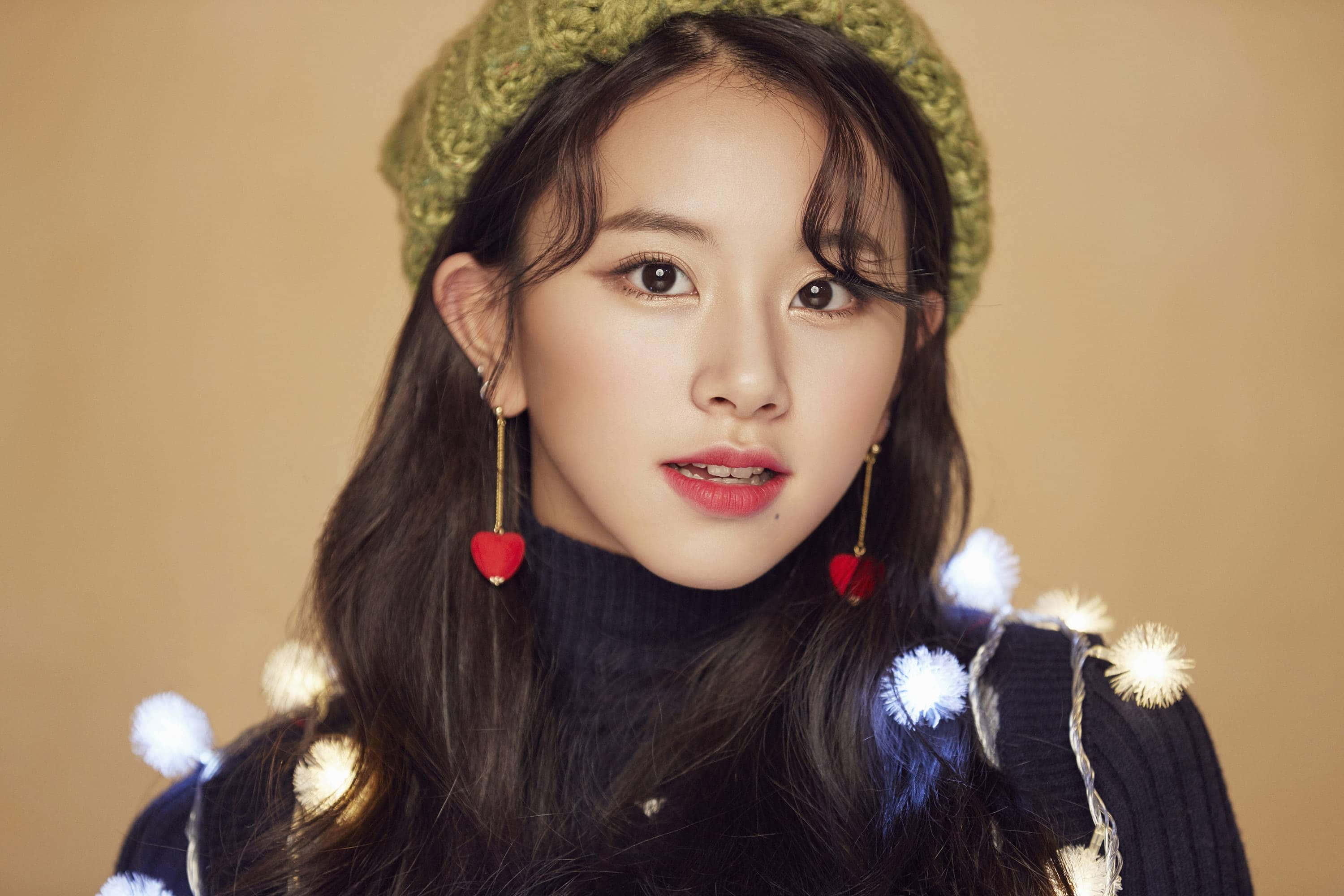twice 桌布 壁紙 — 一位長黑髮的年輕女性，在溫暖的米色背景前，戴著綠色針織貝雷帽，穿著飾有白色絨球和紅色心形耳環的深色毛衣。