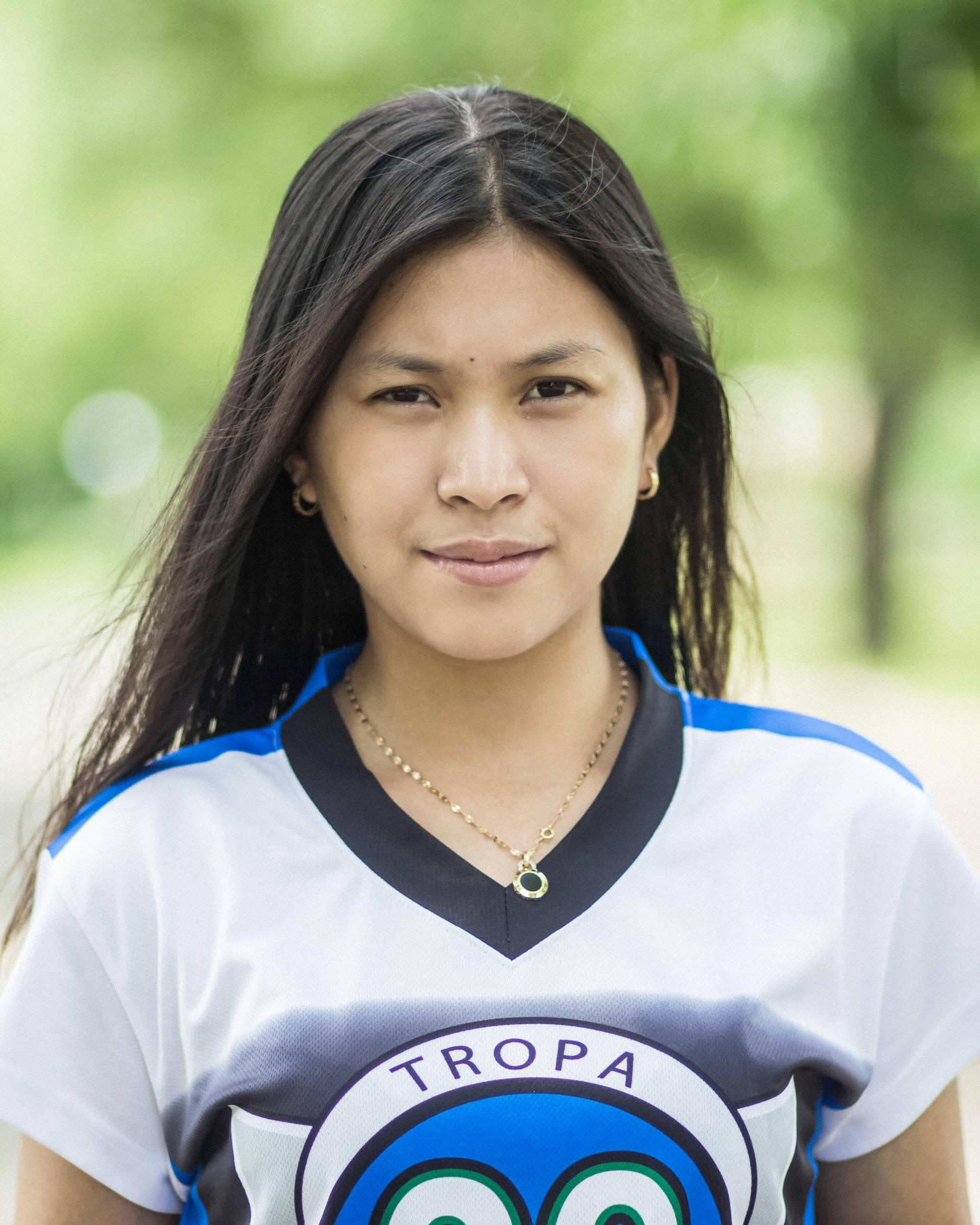 headshot 桌布 壁紙 — 一位長黑髮的年輕女性，穿著帶有 'TROPA' 標誌的白色、藍色和黑色運動球衣，在模糊的綠色公園背景下溫柔微笑。