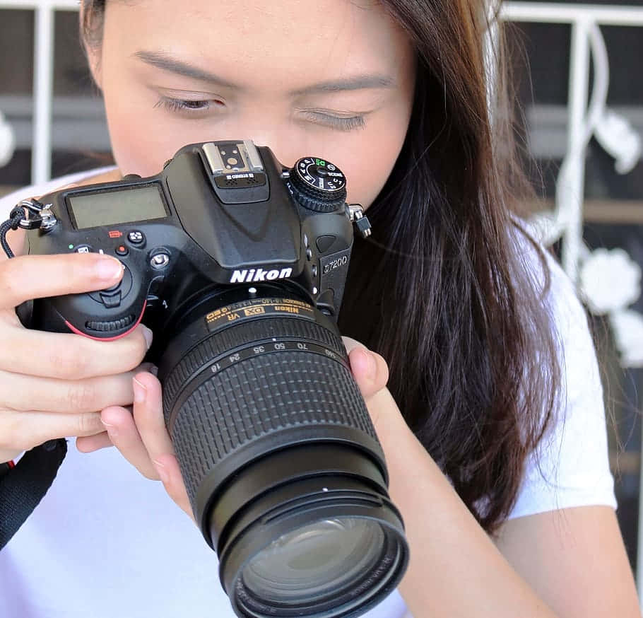 攝影 桌布 壁紙 — 一位長黑髮的年輕女性，穿著白色T恤，將黑色Nikon DSLR相機舉到眼前。