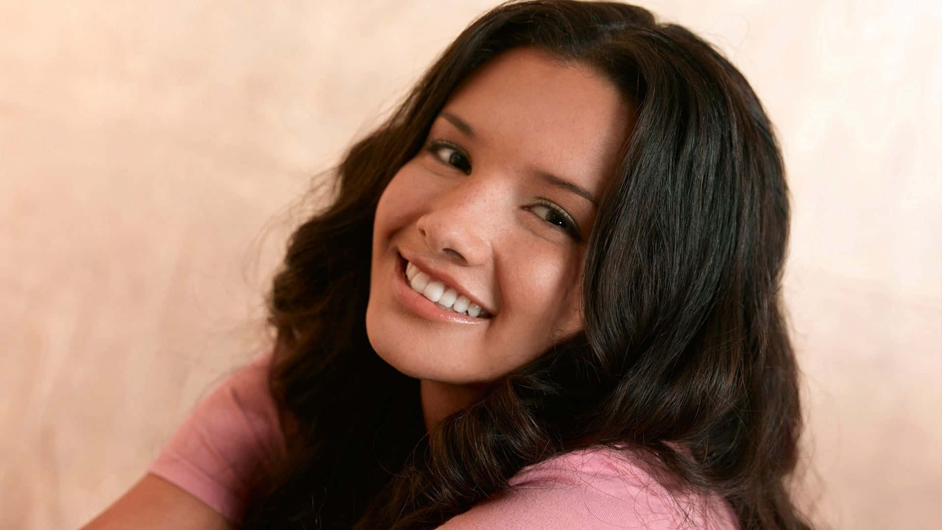 Alice Greczyn 桌布 壁紙 — 一位長黑髮的年輕女性，穿著粉紅色襯衫，在柔和的米色牆壁前溫暖地微笑。