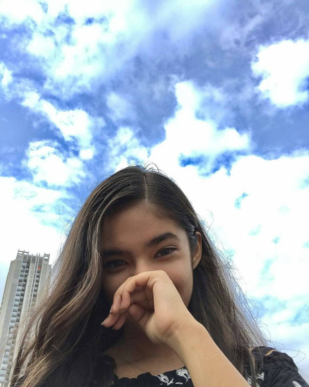 Indian 桌布 壁紙 — 一位長黑髮的年輕女性在戲劇性的藍天和蓬鬆白雲背景下，可見一棟高樓，頑皮地捂住嘴巴。